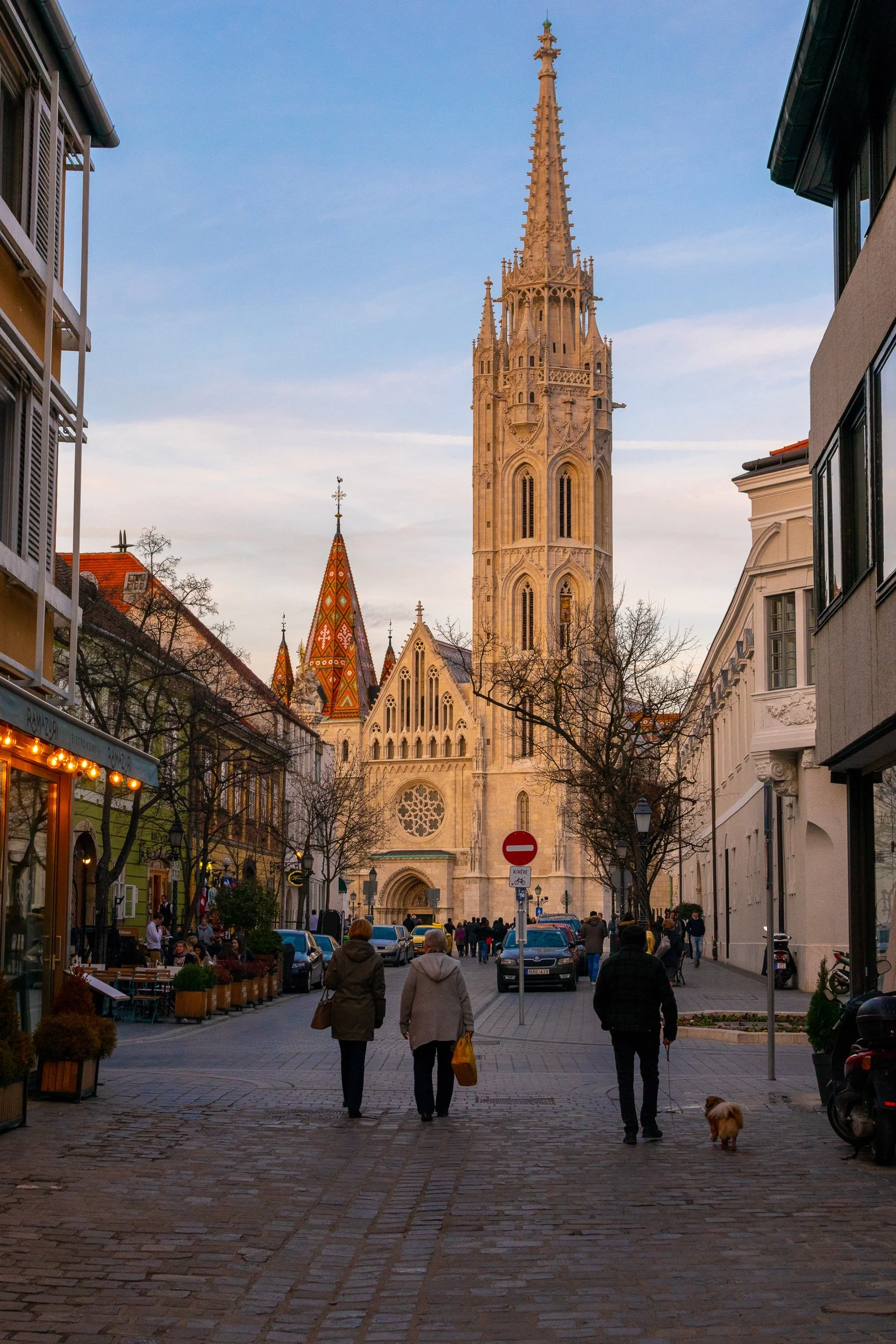 Budapest street scene, Matthias Church in the background, people walking on cobblestones in warm evening light.