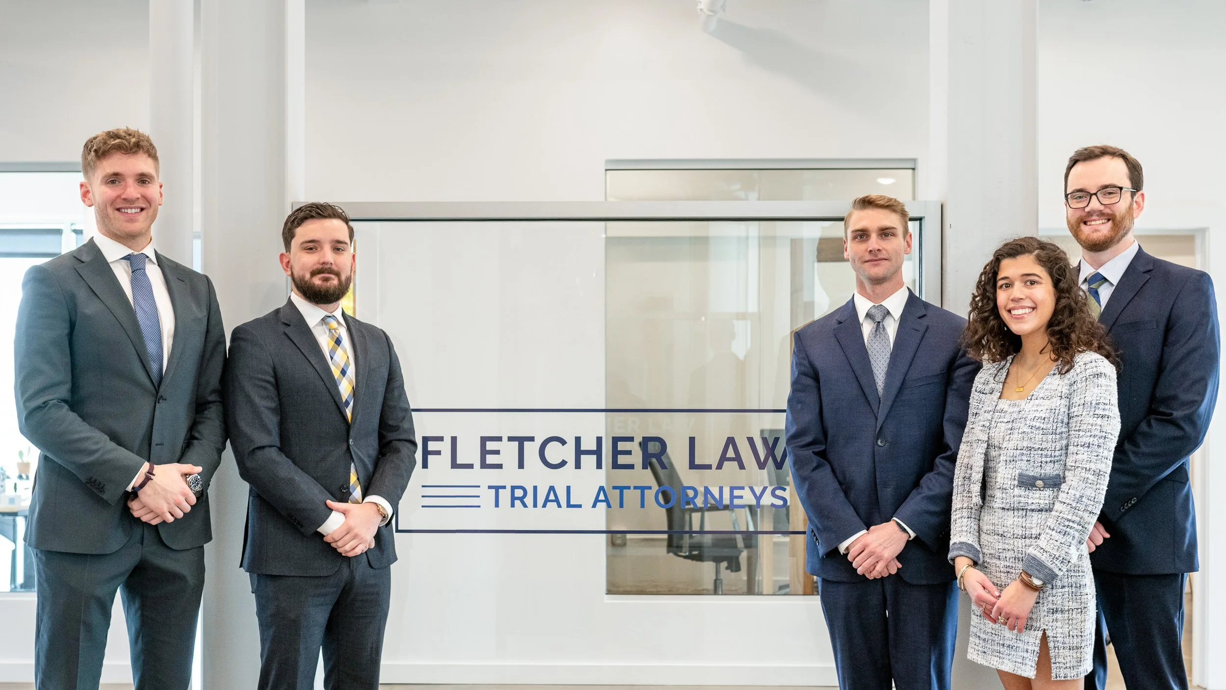 Five attorneys in suits standing in front of a Fletcher Law Trial Attorneys branded glass partition in a modern office

