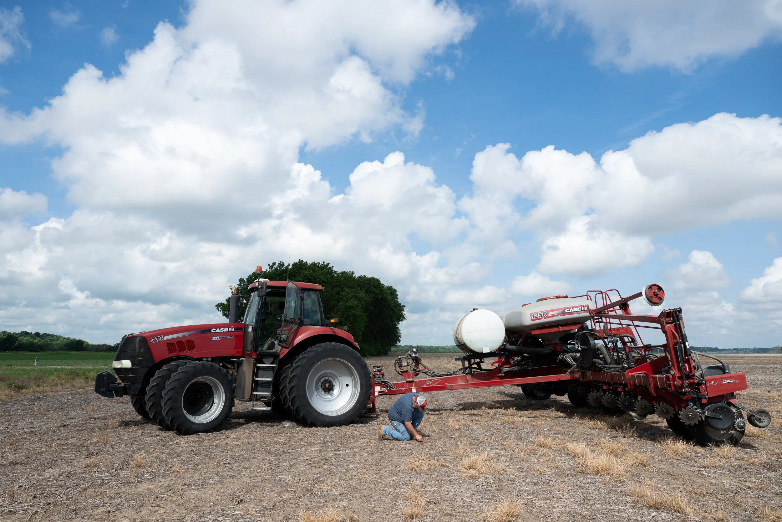 Farmer servicing a red tractor and air seeder in a cultivated field beneath a blue sky.