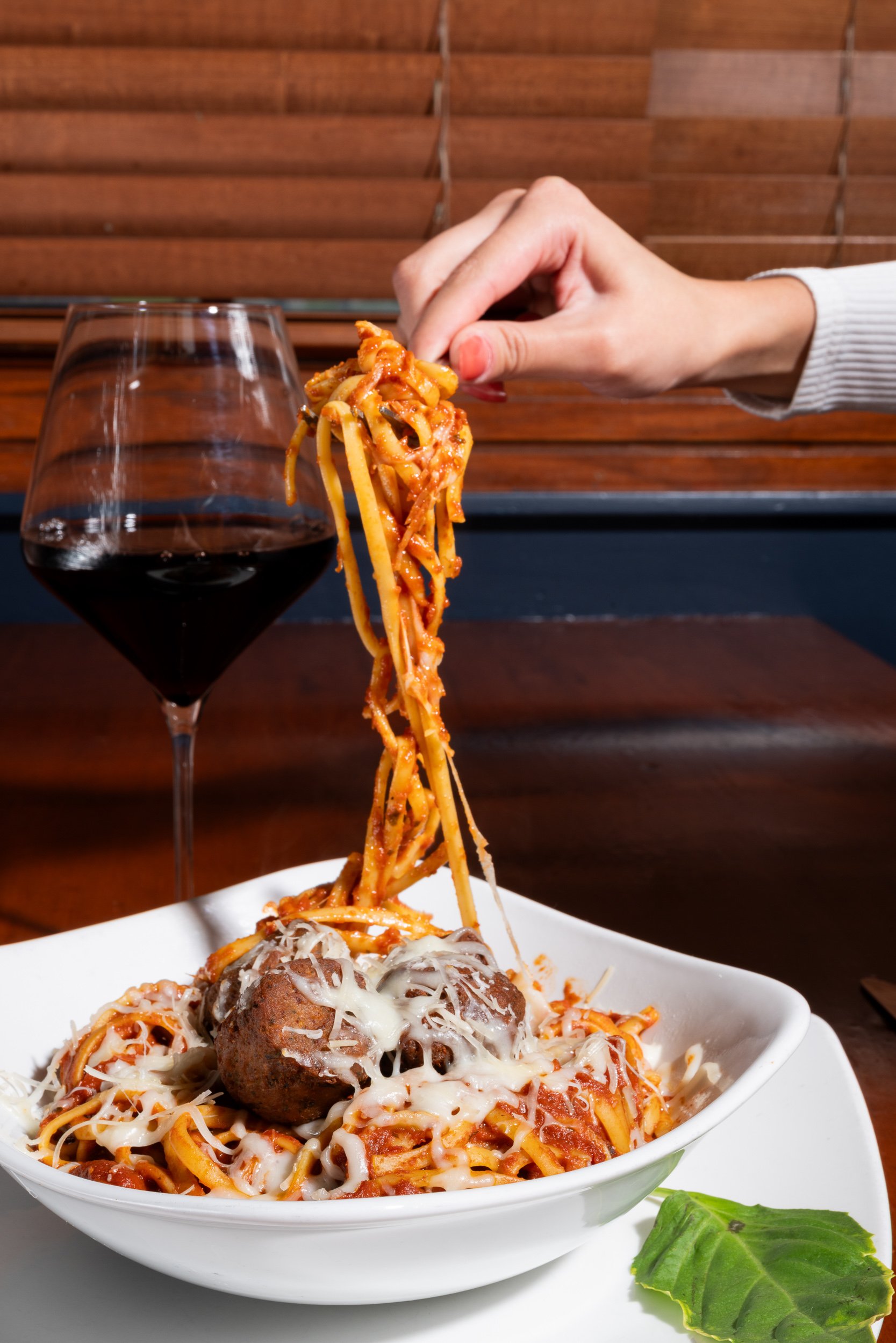Hand twirling spaghetti with meatballs and red wine glass, restaurant dining experience photography

