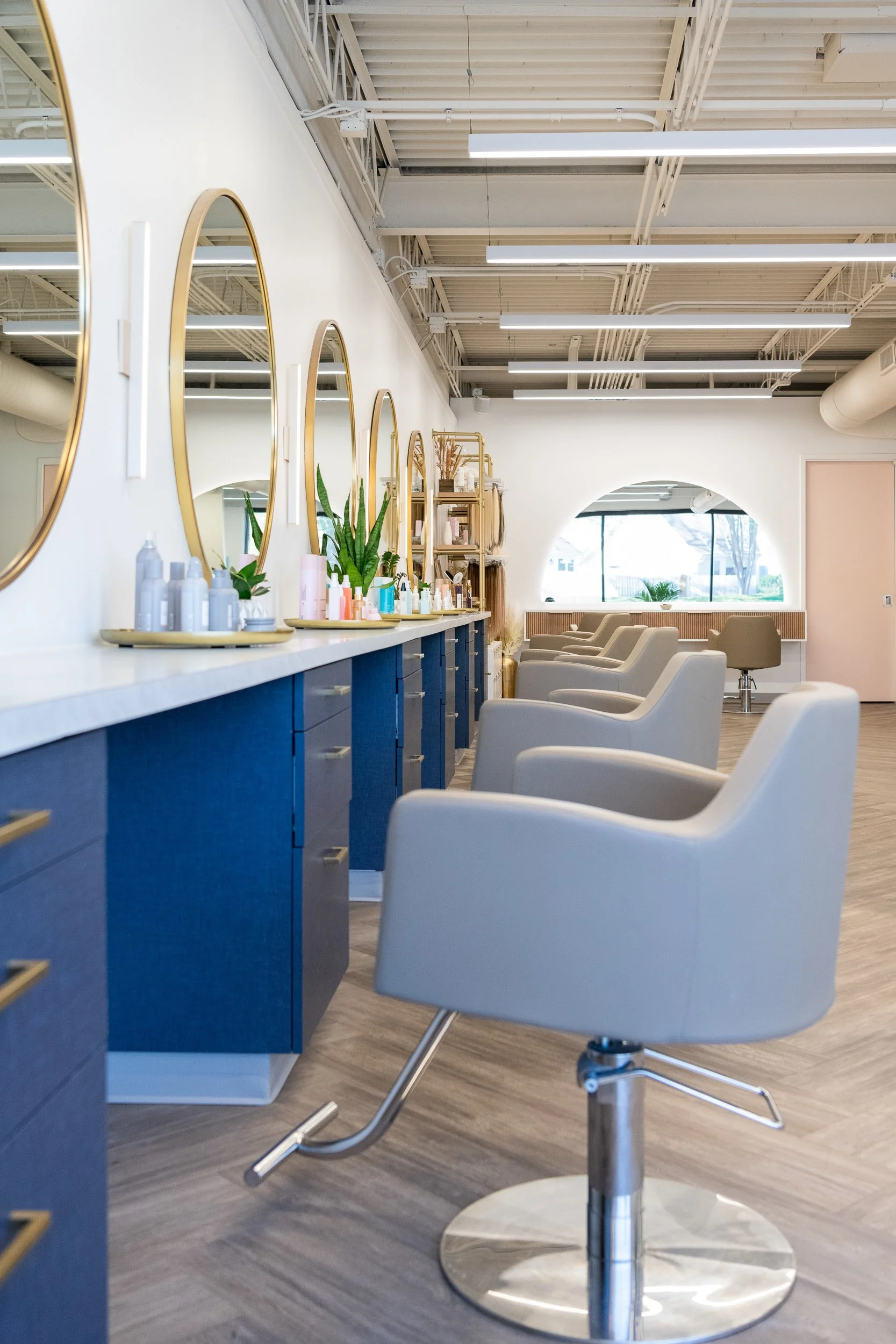 Row of styling chairs and gold oval mirrors along a blue cabinet in a modern hair salon