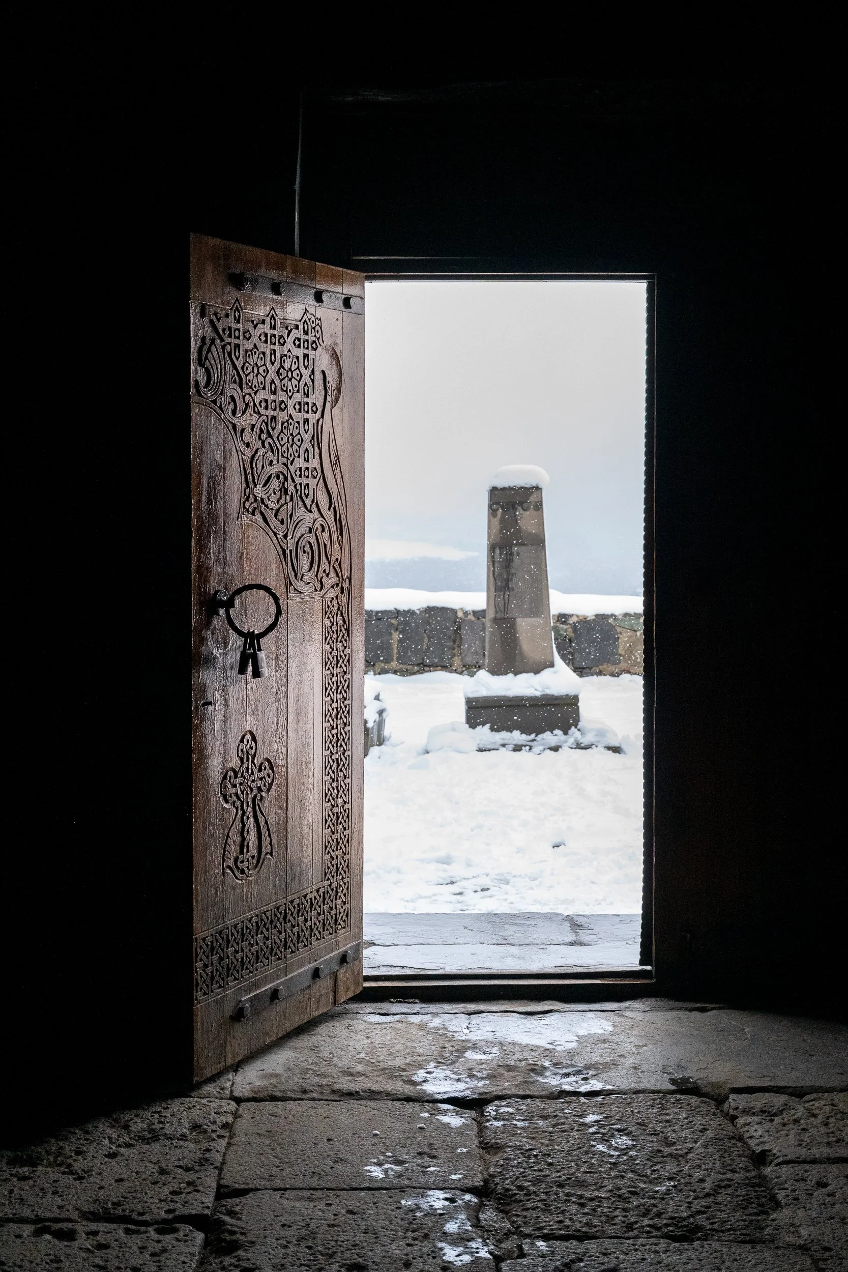 An intricately carved wooden doorway opens from darkness into a snow-covered monastery courtyard, inviting the viewer to step from shelter into silence.