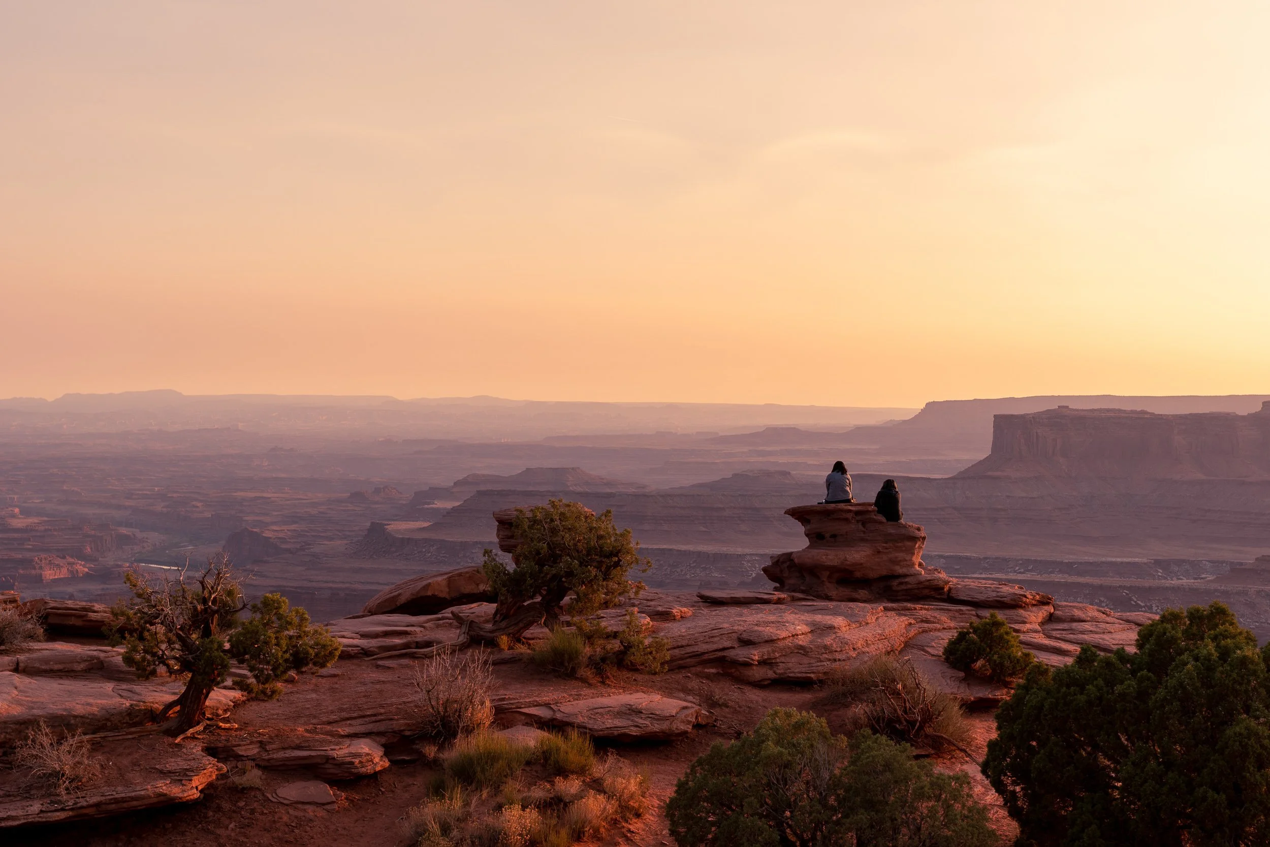  Dead Horse Point State Park, Utah at sunset — two figures sitting on a rock formation, warm orange canyon light, mesa stretching to the horizon.