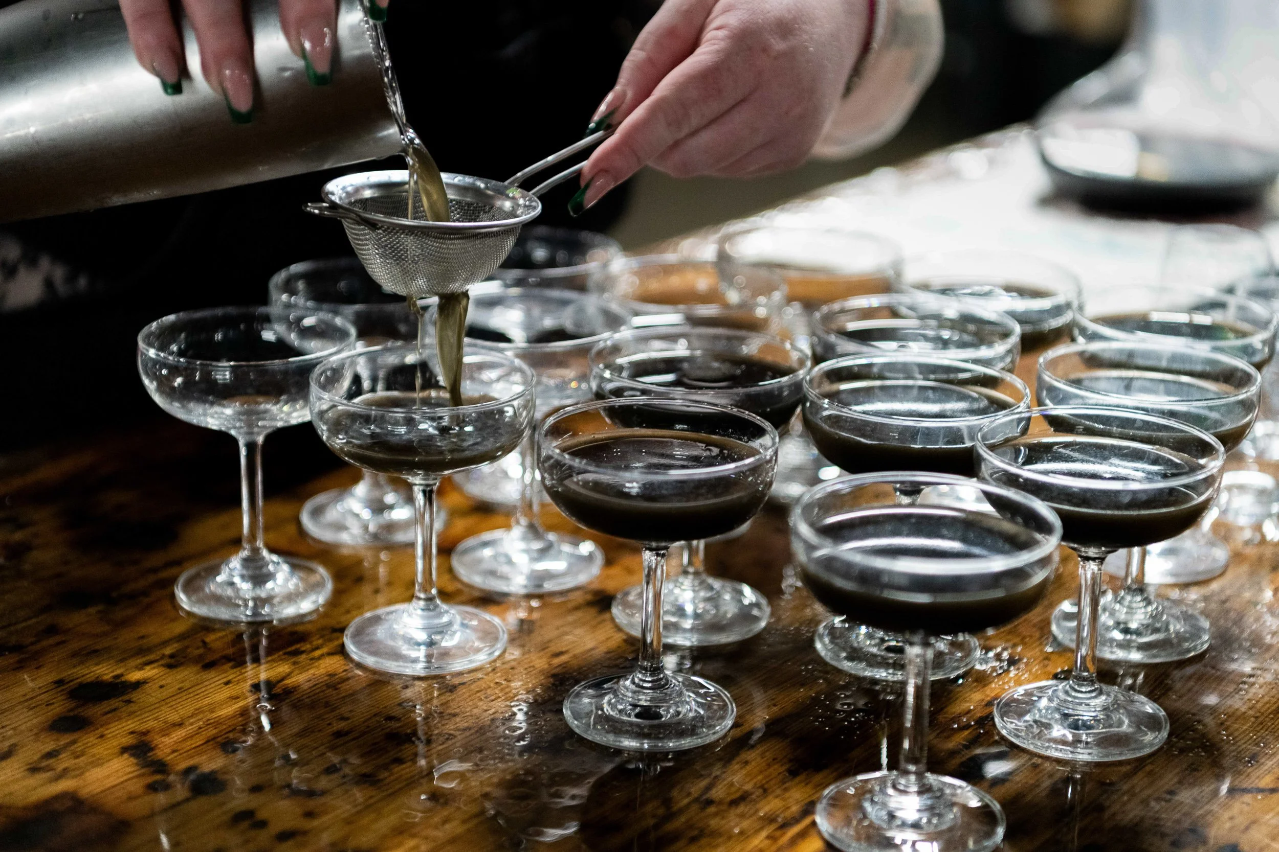 Bartender pouring espresso martinis into a row of coupe glasses, bar service photography

