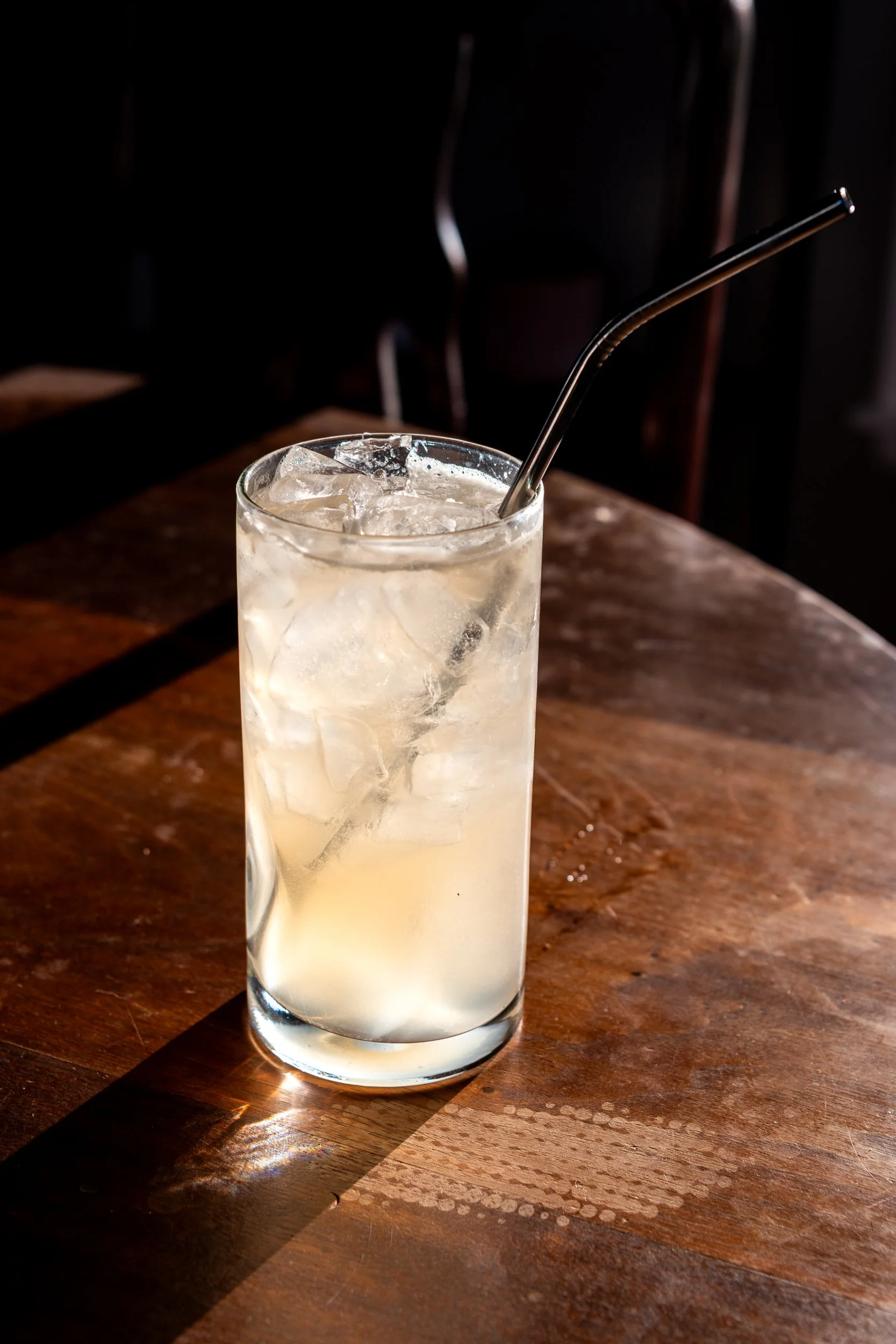 Clear highball cocktail with ice and metal straw on wood bar, restaurant beverage photography
