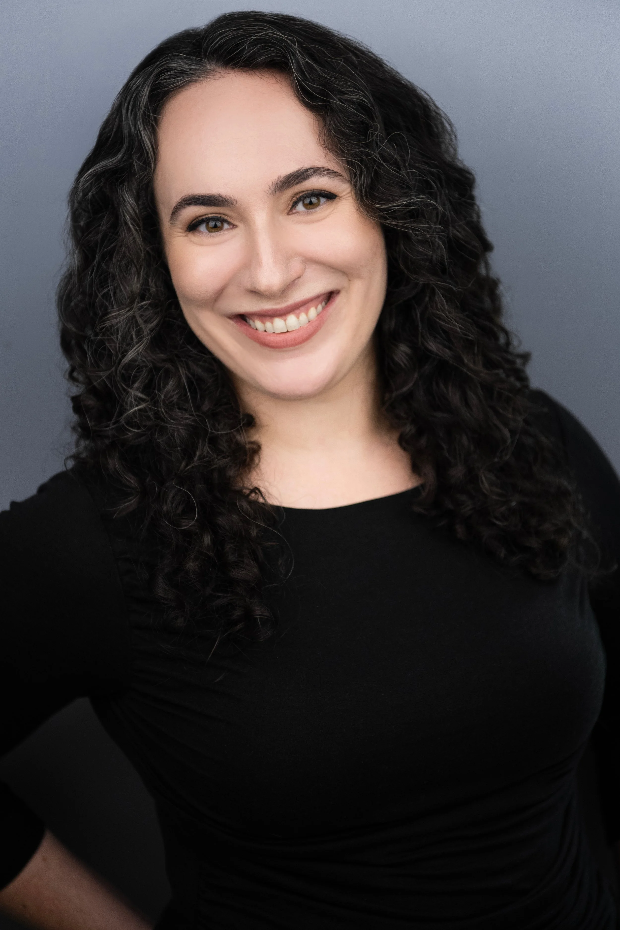 Close-up of a woman with curly dark hair, smiling, wearing a black top, against a gray background.