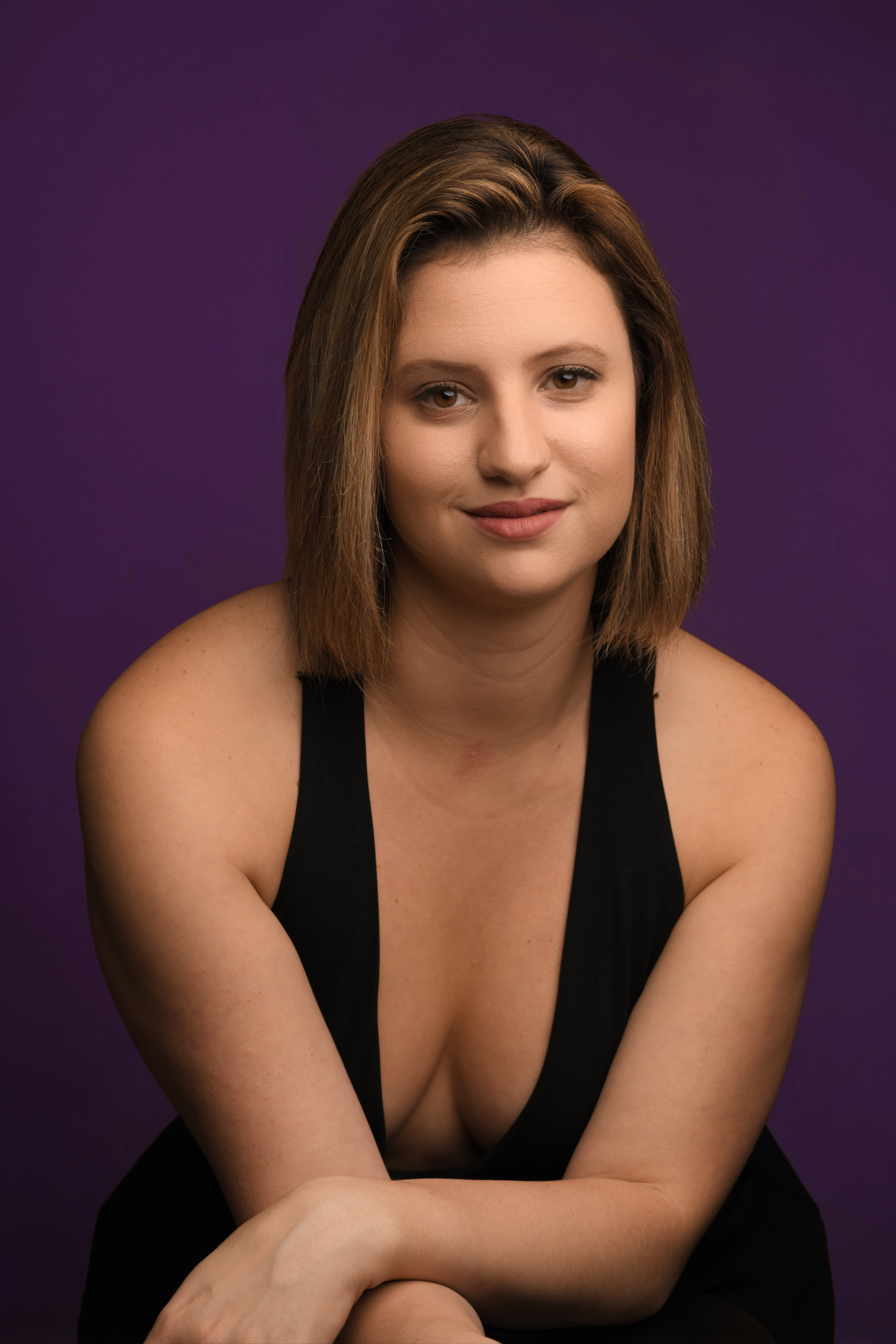A woman with shoulder-length brown hair and brown eyes, smiling slightly, wearing a black sleeveless top, sitting with her arms crossed in front of her on a purple background.
