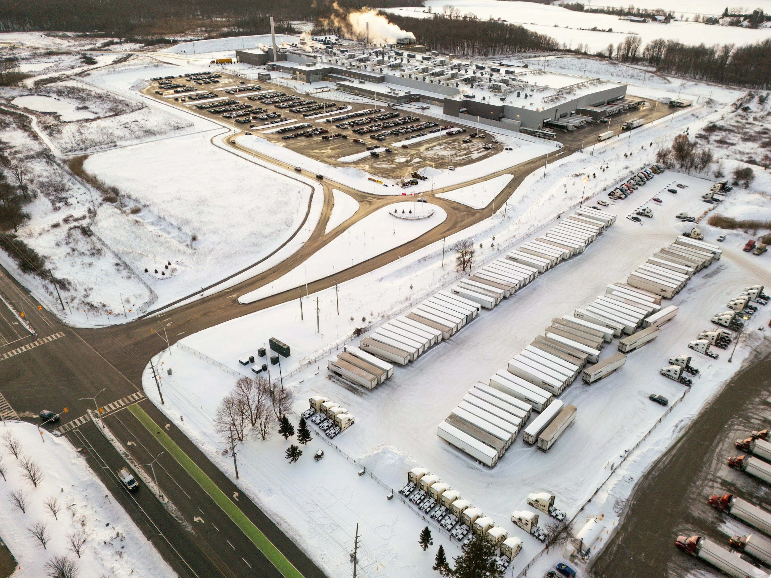 Industrial landscaping services. Industrial snow removal services. Aerial view of a large warehouse surrounded by snow-covered landscape and multiple parking lots filled with vehicles and trucks.