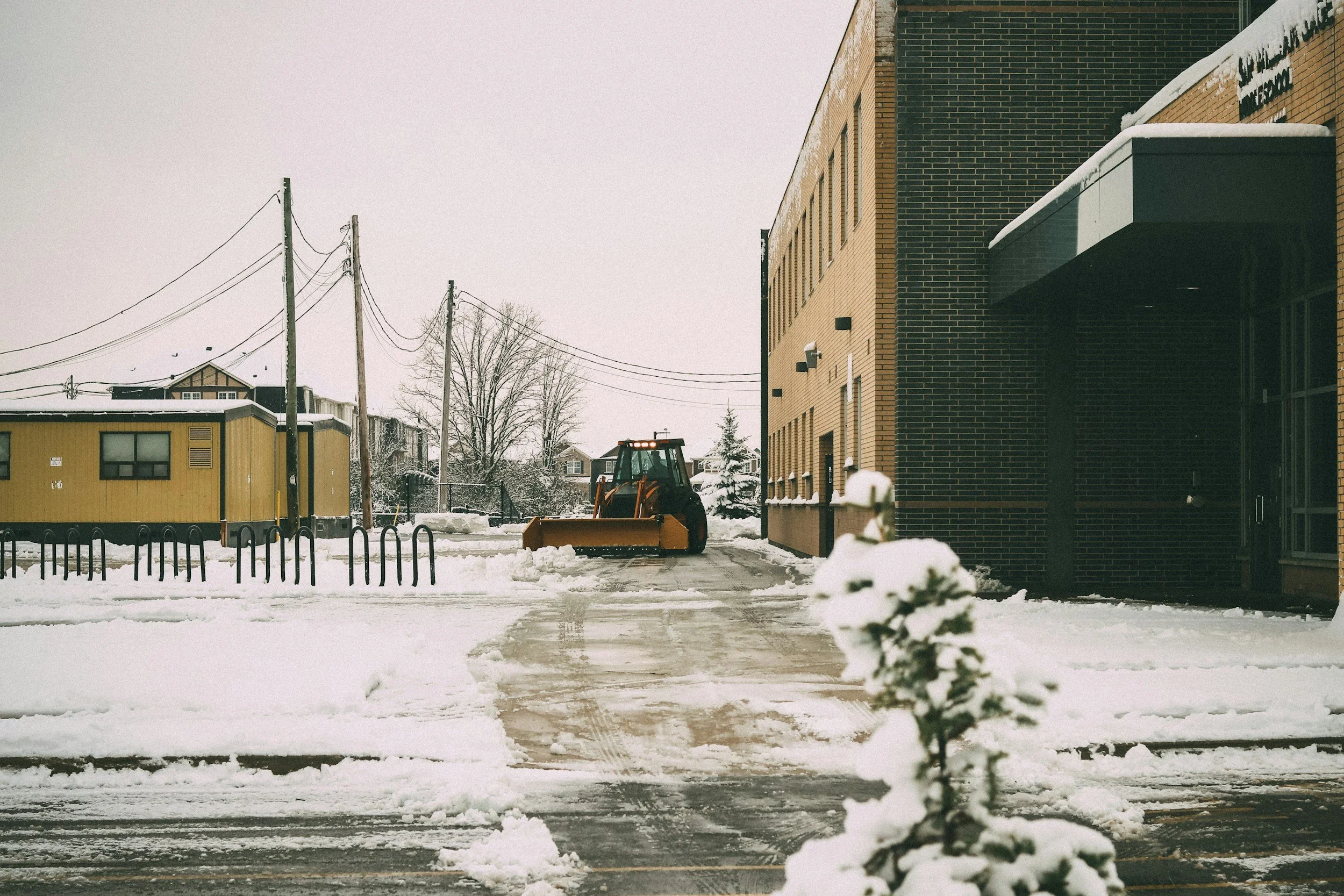 Industrial landscaping services. Industrial snow removal services. Snow-covered street with a tractor plowing snow, modern buildings, leafless trees, utility poles with wires, cloudy sky, and snow on the ground. Industrial Facilities Management