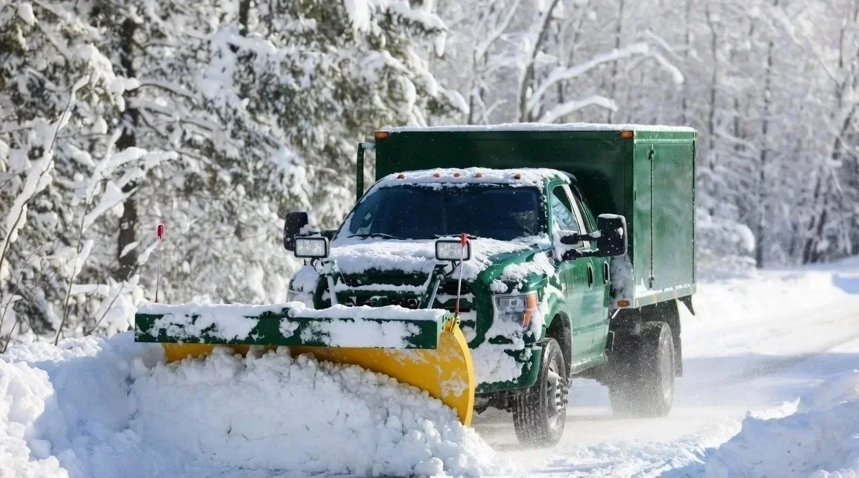 Professional Snow Removal Services. Commercia Snow Removal Services. Green pickup truck equipped with a snow plow clearing snow from a road in a snowy landscape with trees covered in snow.