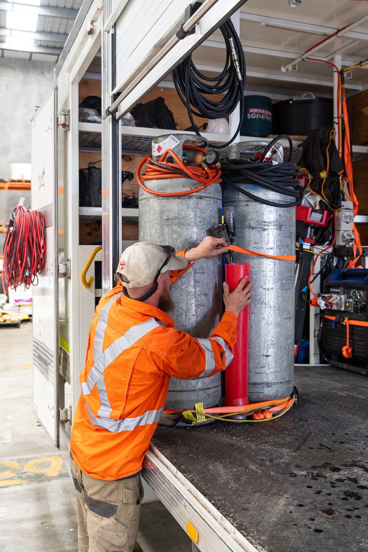 A worker in an orange safety jacket and beige cap is securing a red object to a large metal tank inside a truck, with other equipment and tools visible in the background.