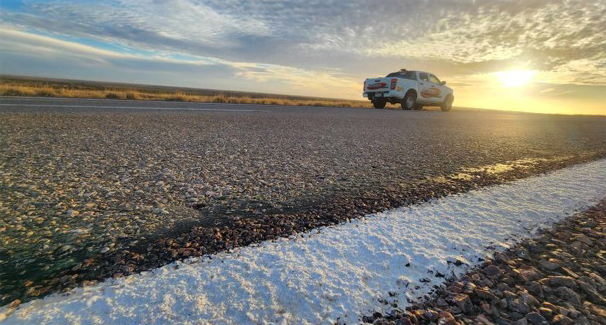 A white ute driving on a rural road at sunset, viewed from a low angle near the edge of the road with a white line marking the shoulder.