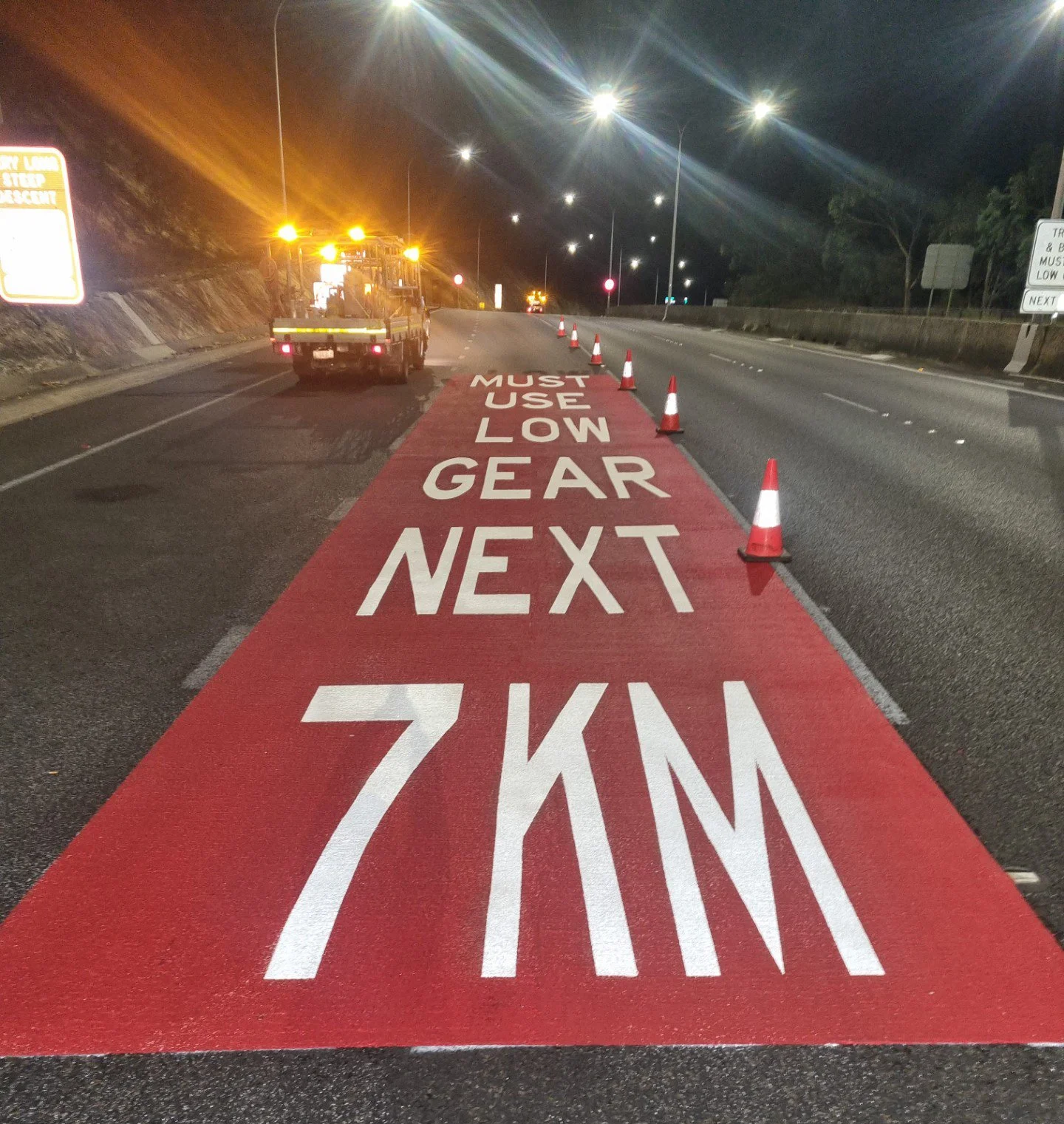 Nighttime scene of a road with orange traffic cones and a red painted lane that reads: "MUST USE LOW GEAR NEXT 7KM." A vehicle with flashing lights is parked nearby. There are illuminated streetlights and signs along the side of the road.