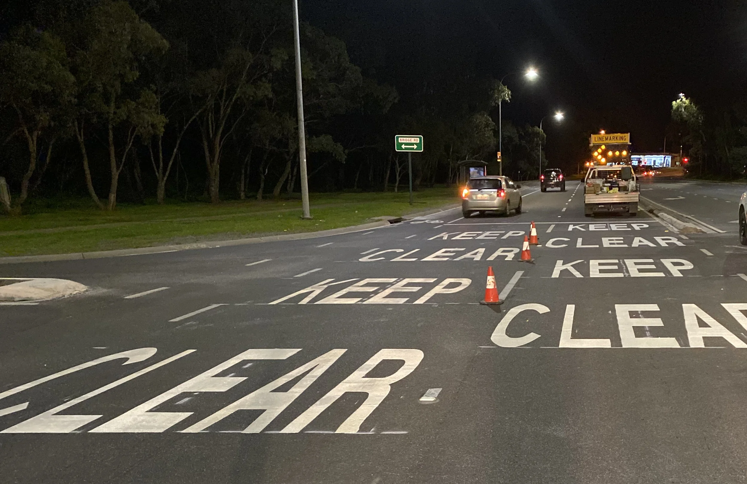 Nighttime scene showing a road with multiple cars, a lane closure with cone markers, a sign indicating lane merging, and a green sign for Bridge Road. The area is surrounded by trees and illuminated by streetlights.