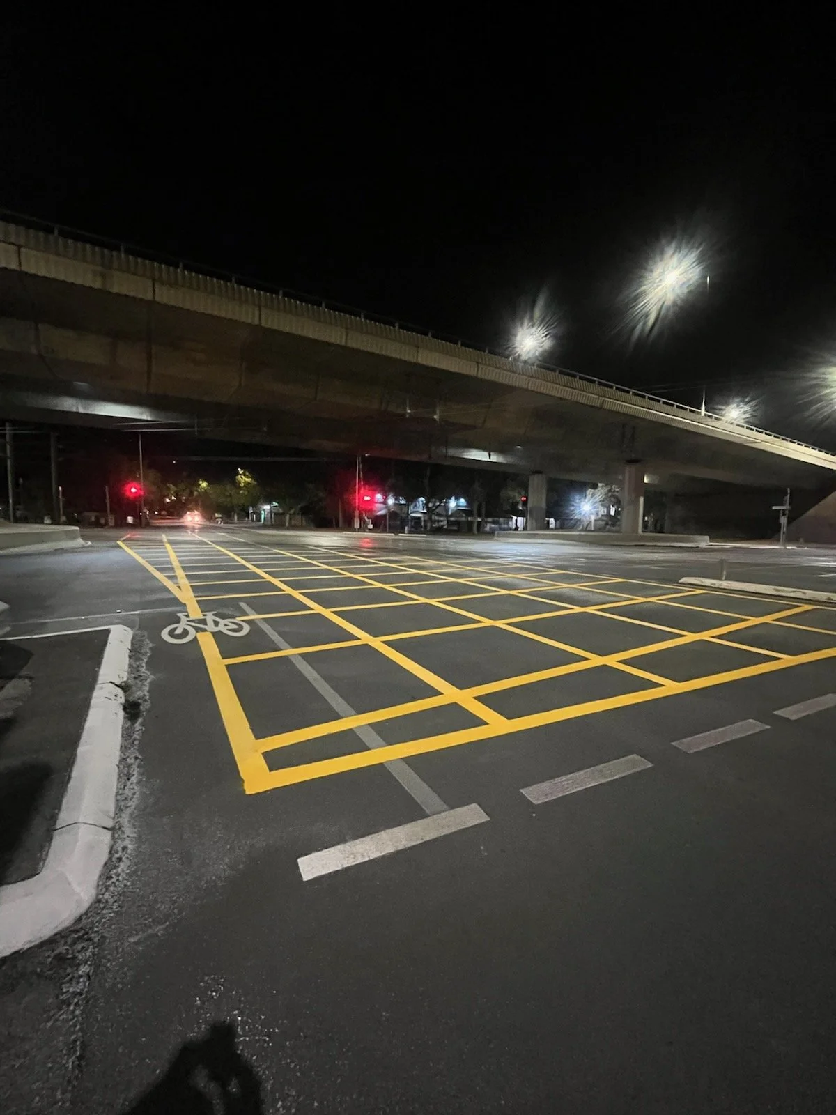 Empty parking lot at night with yellow grid lines, a designated bicycle lane, under a bridge with streetlights, and traffic signals in the background.