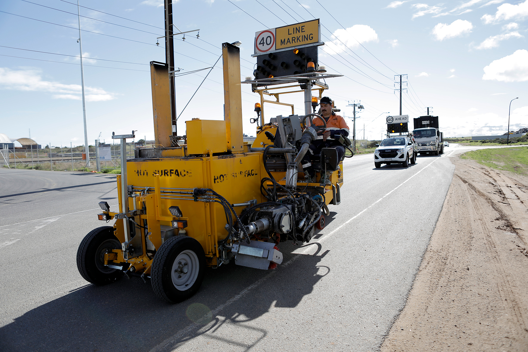 Road construction crew using a yellow machine to mark lane lines on a road while cars follow behind with a sign indicating 40 mph speed limit and ahead warning.
