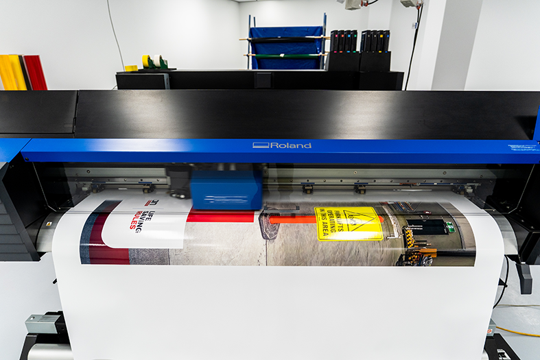 A large Roland printing machine printing a banner with warning signs and text.