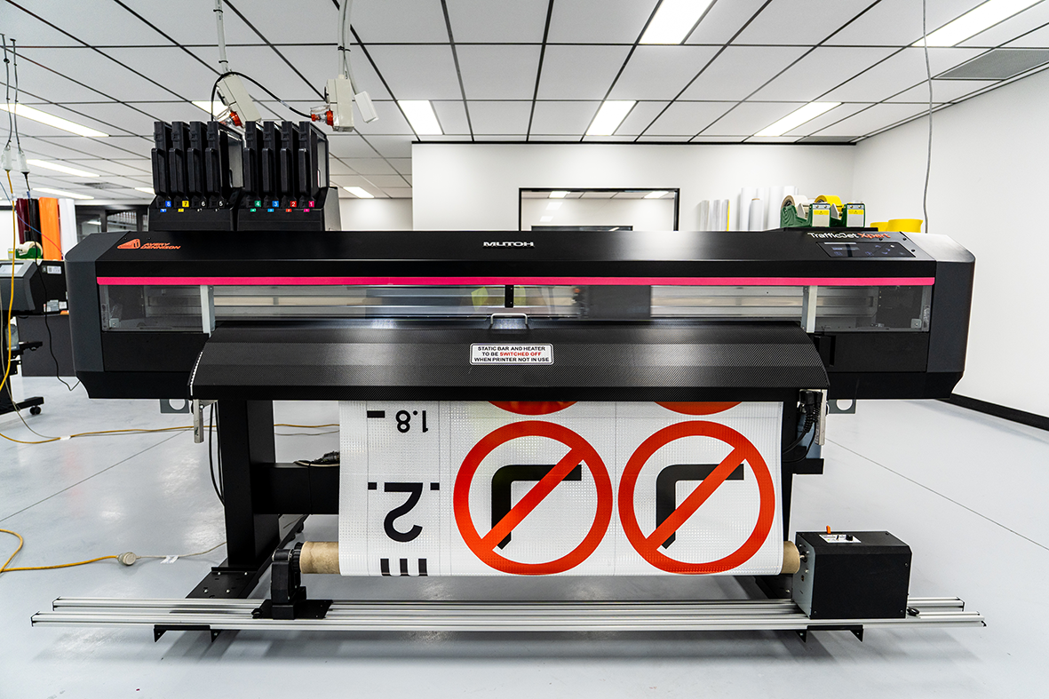 Large digital printer printing a sign with no parking symbols on a roll of material in a well-lit room.