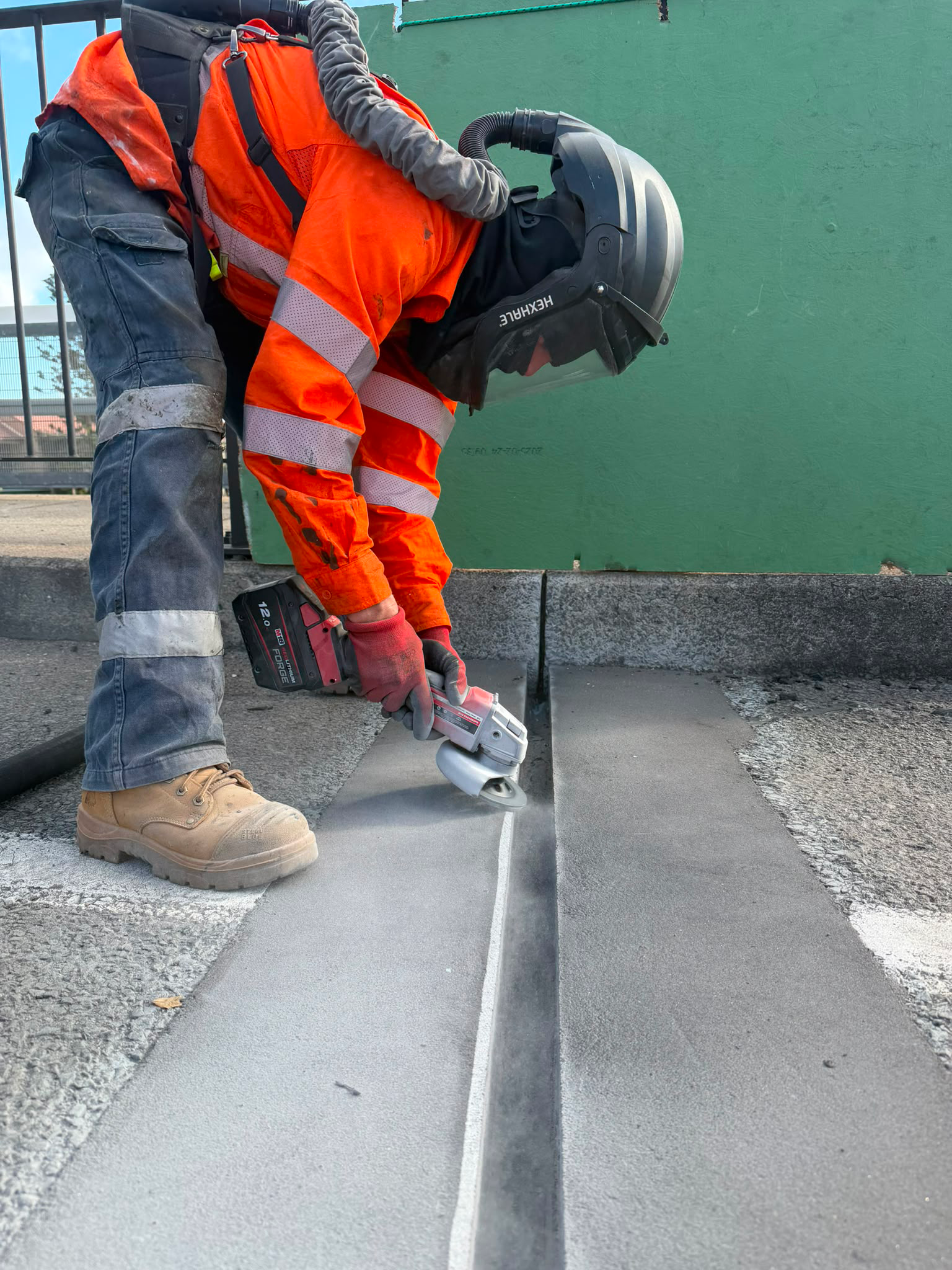 Worker in orange safety vest and helmet using a power tool to pour concrete onto a sidewalk.