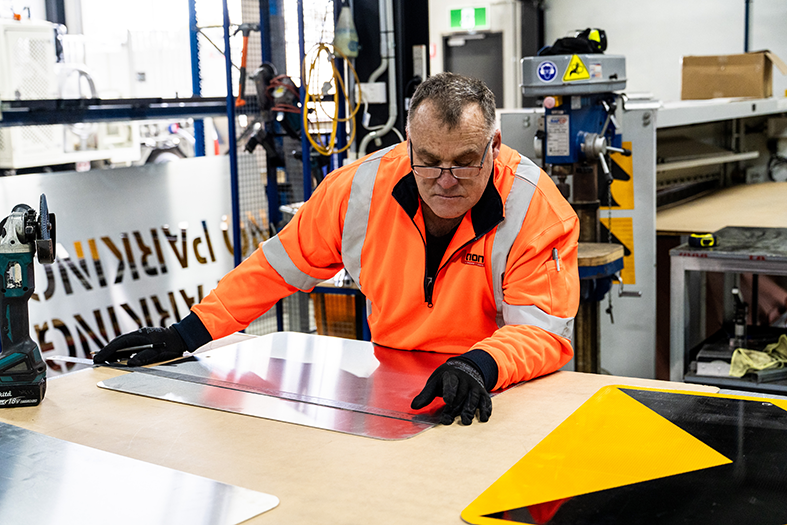 A worker in an orange safety jacket and black gloves measures a reflective sheet at a workbench in a sign manufacturing workshop.