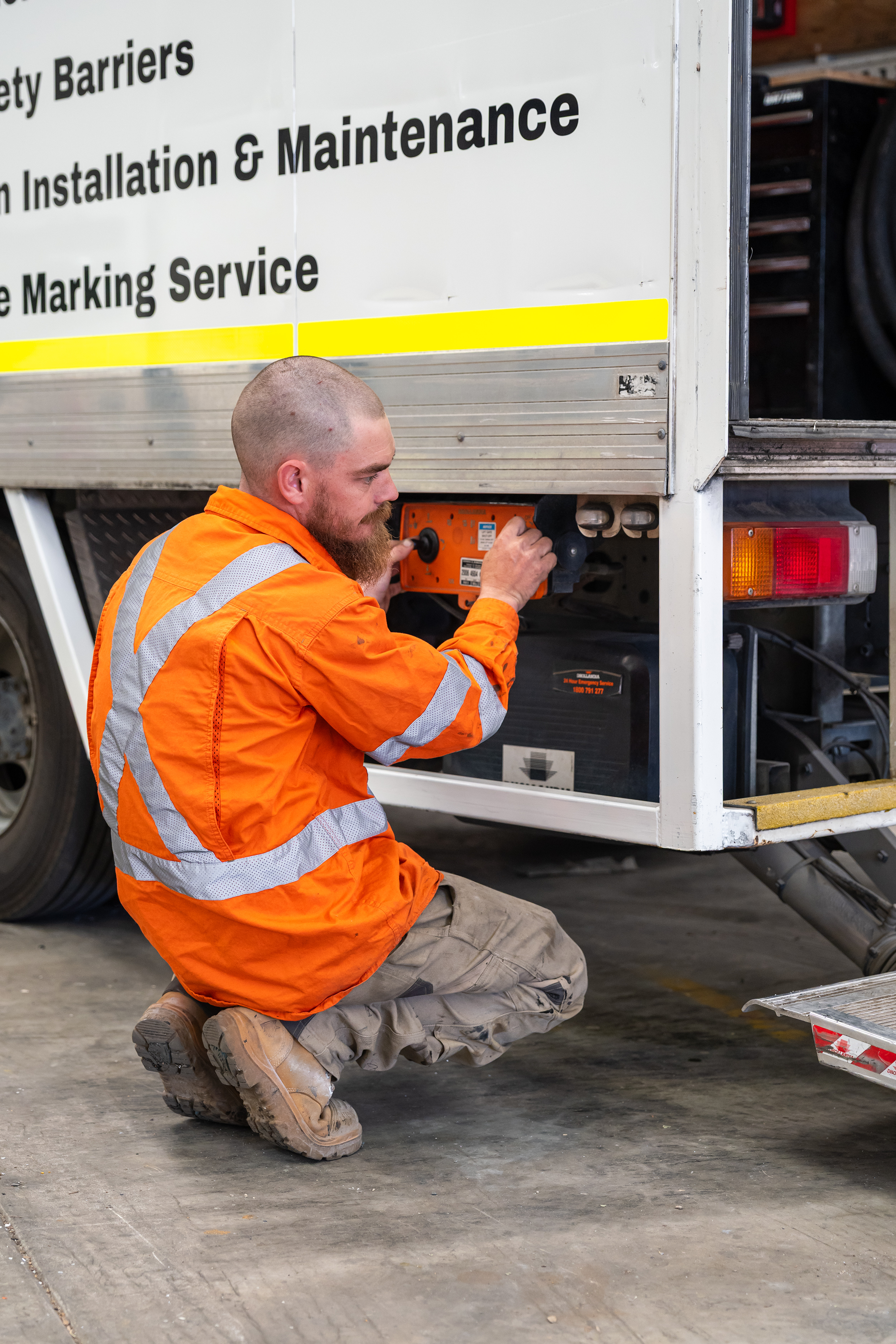 A worker in orange safety gear kneeling on the floor and operating equipment on a service truck with a sign that reads 'Safety Barriers, Installation & Maintenance, Marking Service'.