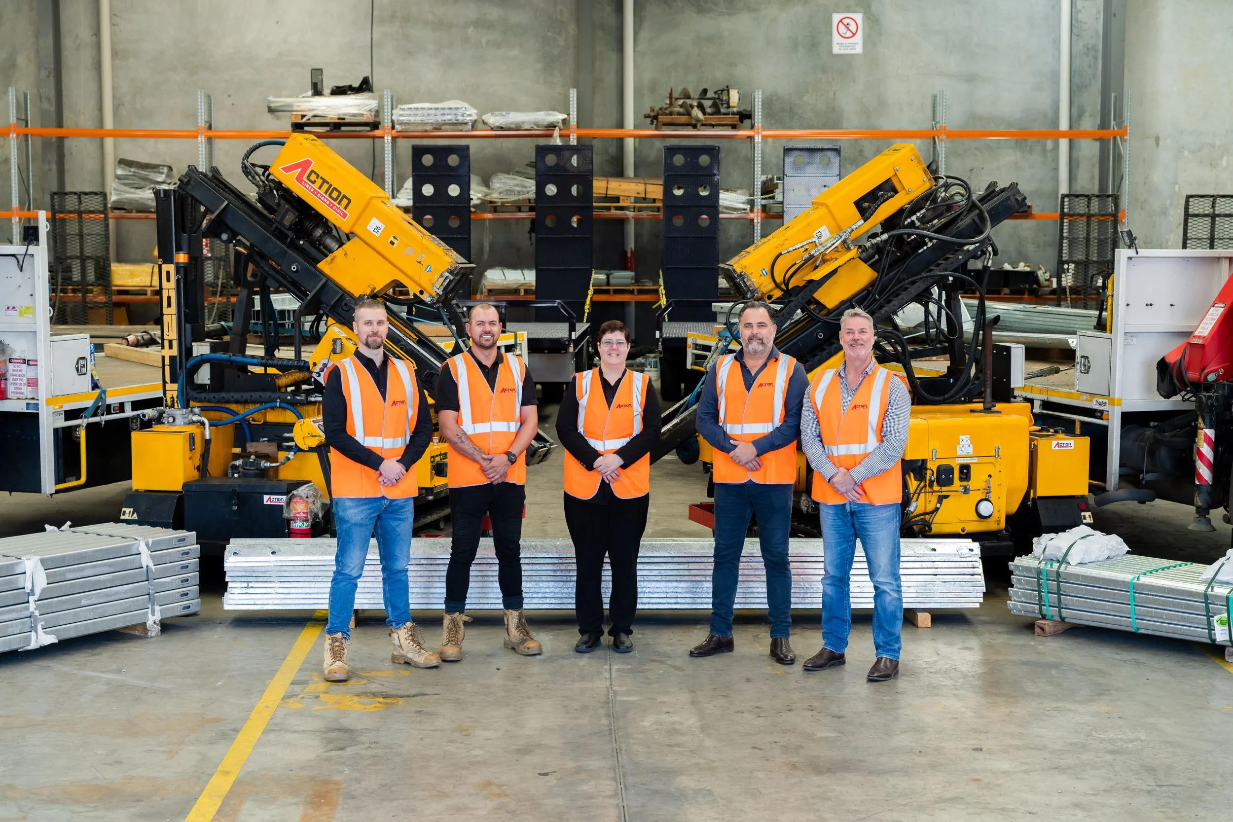 Five workers in orange safety vests and casual clothing standing in front of industrial machinery in a warehouse.
