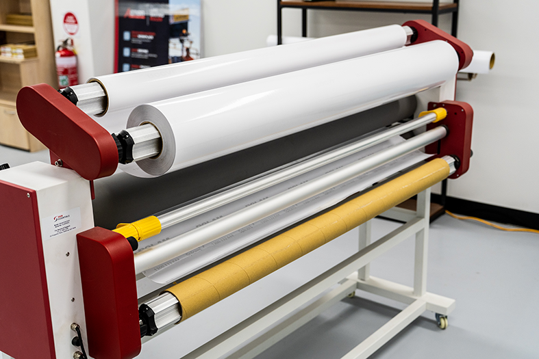 Industrial laminating machine with white and yellow rolls of material in a workspace.