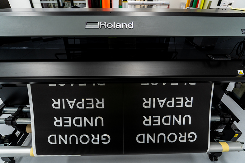 A Roland UV printer printing upside-down text that says 'GROUND UNDER REPAIR'.