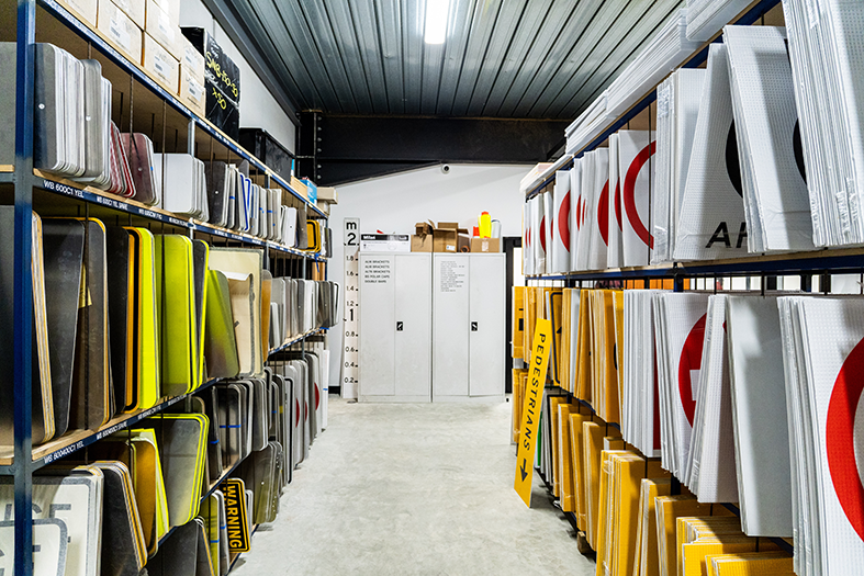 Indoor storage room with metal shelves stocked with traffic signs, lane markers, and reflective panels.