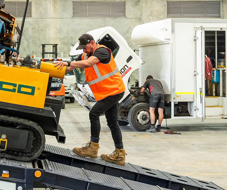 A man wearing a black cap, orange safety vest, black pants, and brown boots is balancing on a metal ramp and working on a piece of heavy machinery indoors. In the background, another person is working on the side of a white box truck, which is parked