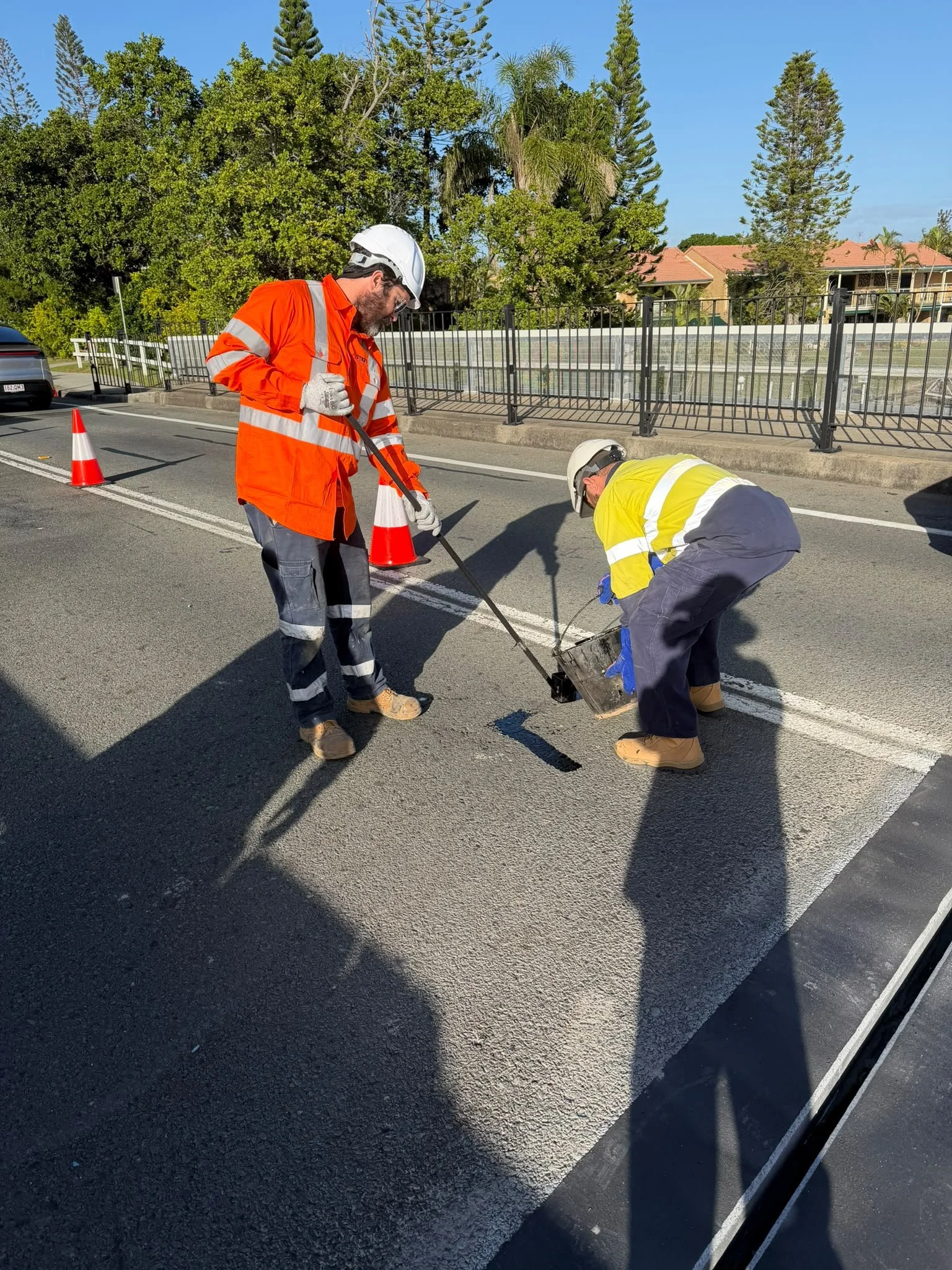 Two construction workers wearing safety gear are working on a street. One is in an orange safety jacket and the other in a yellow safety jacket. They are using tools near a freshly painted white line on the road. Traffic cones are set around them.