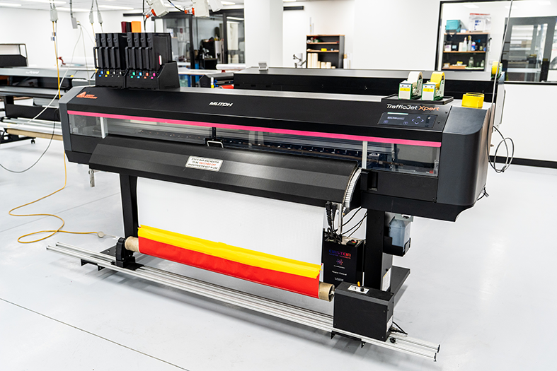 Large industrial printer in a clean, modern workspace, printing a roll of material with yellow and red stripes.