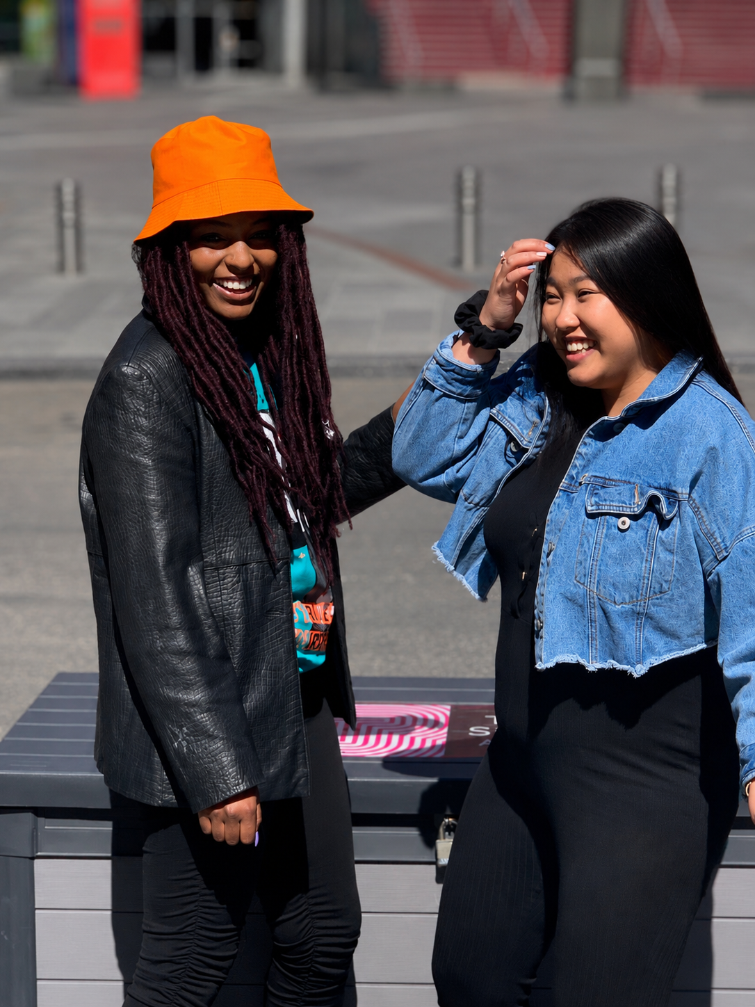 Two women smiling and interacting outdoors in a city, one with purple dreadlocks wearing a bright orange hat and a black leather jacket, the other with long black hair in a denim jacket, with a gray table in the background.