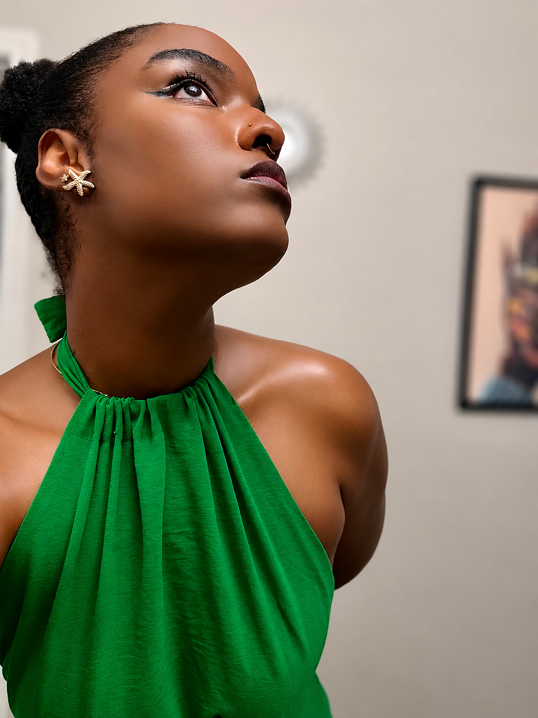 A woman with dark skin wearing a green halter dress, gold starfish earrings, and a nose piercing, looking upward with an expression of confidence or contemplation, indoors with a blurred background.