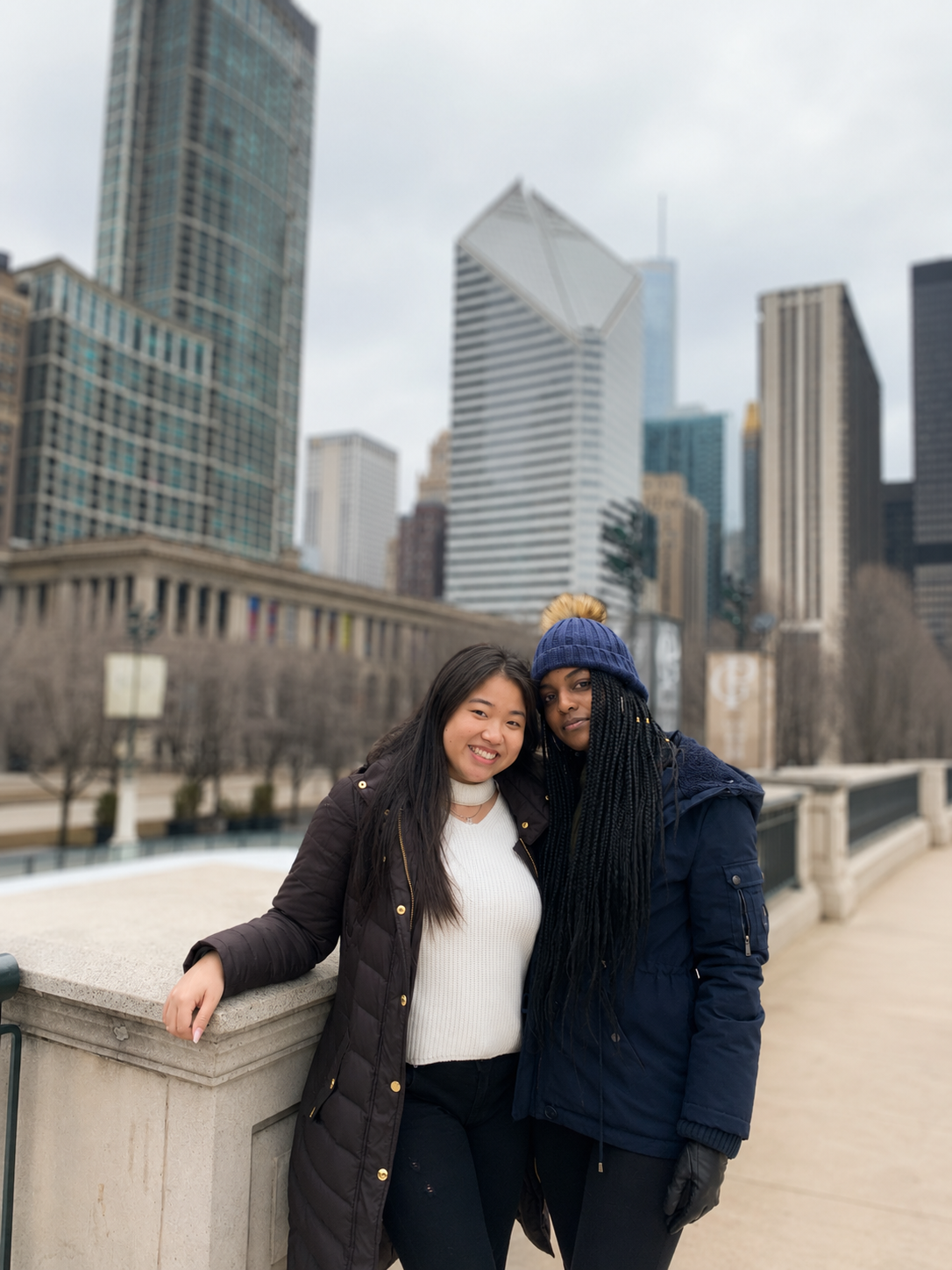 Two women standing close together near a stone railing in an urban park with skyscrapers behind them, one smiling and one with a neutral expression, dressed warmly for cold weather.
