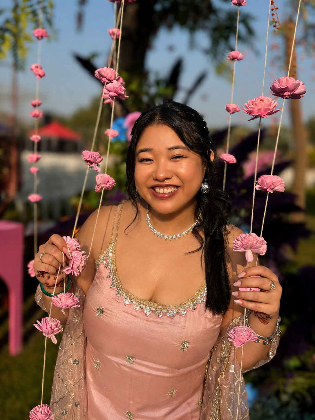 A woman smiling outdoors holding a pink floral decoration, wearing a pink dress with gold embroidery, jewelry, and earrings, with trees and a blue sky in the background.