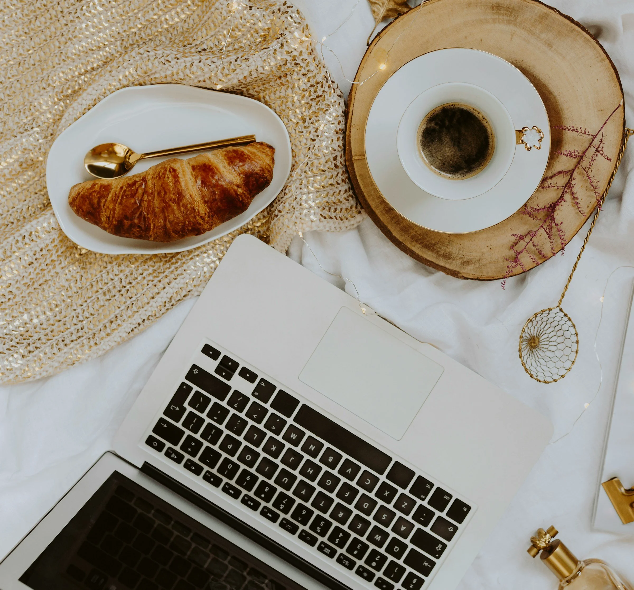 A flat lay of a workspace featuring a MacBook laptop, a white plate with a croissant and a gold spoon, a cup of black coffee on a white saucer, decorative gold accents, a textured beige and gold fabric, fairy lights, and crystal-like ornaments.