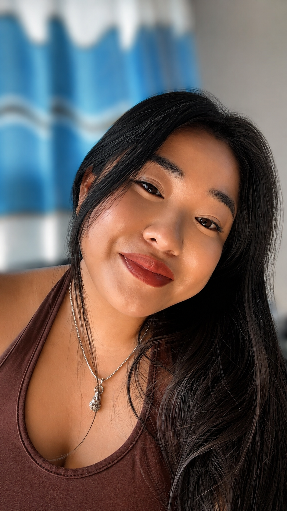 A close-up portrait of a woman with long black hair, wearing a brown top and a silver necklace, smiling softly at the camera.