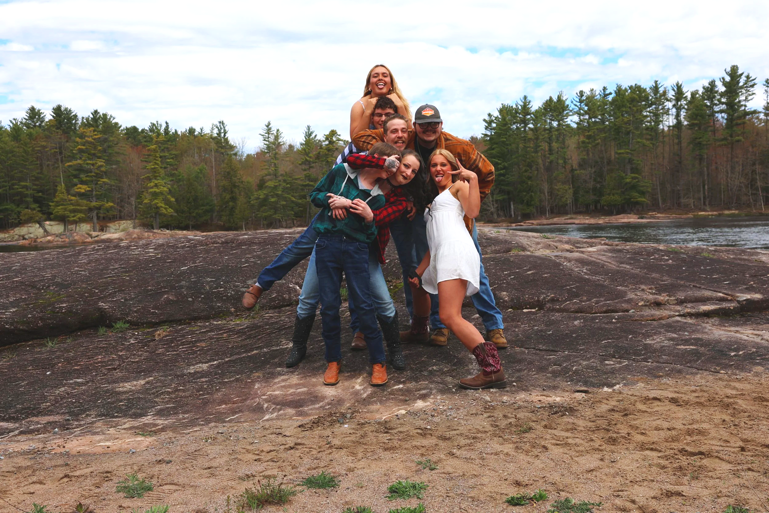 Group of friends posing cheerfully on a large rock by a body of water, with a forest of green trees in the background and a partly cloudy sky.