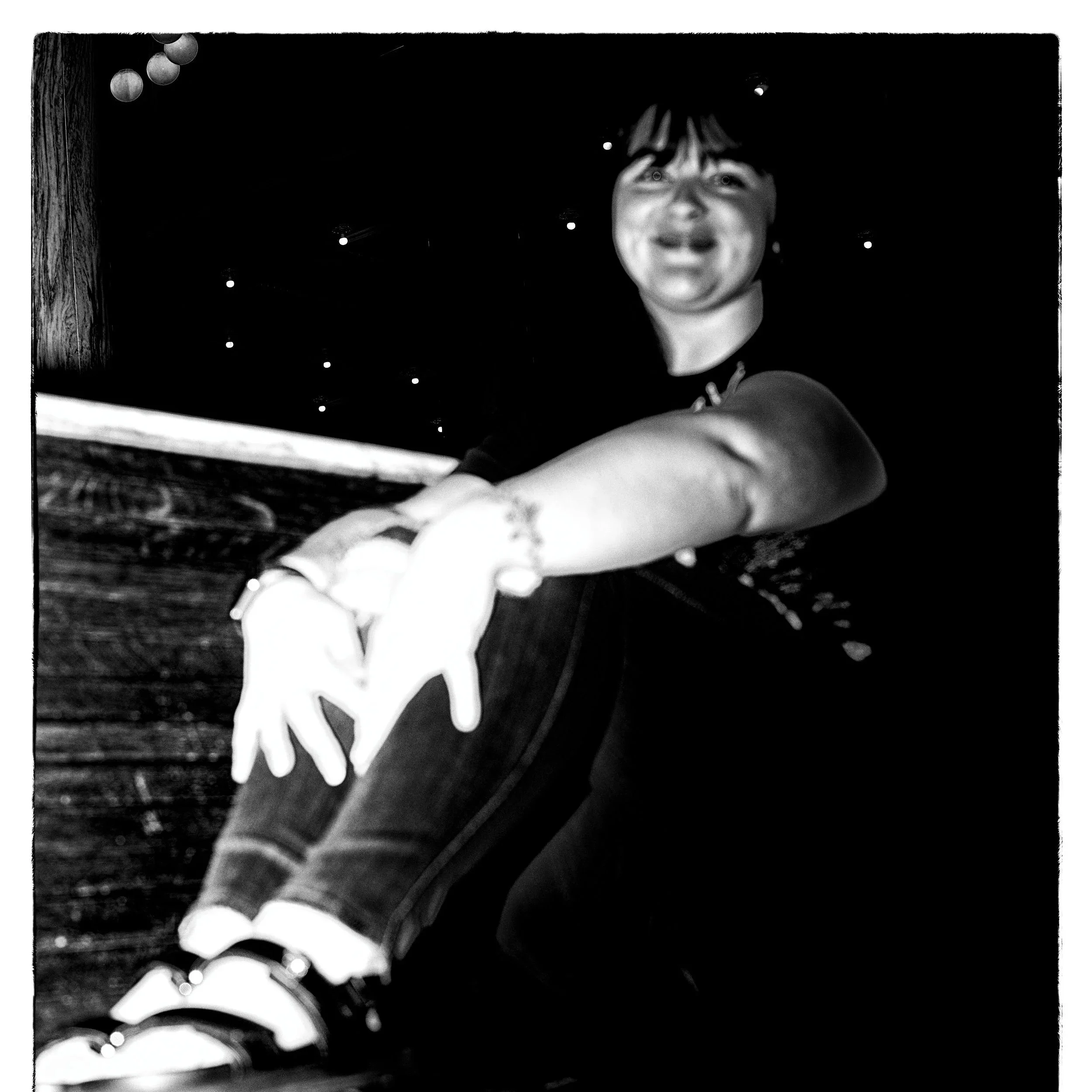 Black and white photo of a smiling young person reaching out with their hand, wearing a short-sleeved shirt and sneakers, set against a dark background with small bright dots and a wooden surface.