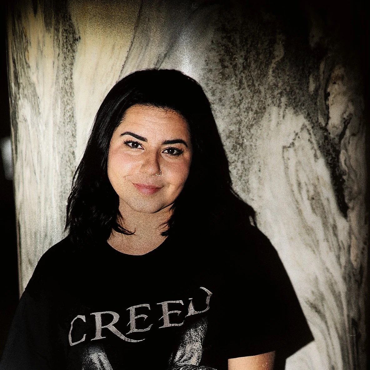 A young woman with black hair, wearing a black Creepx shirt and light blue ripped jeans, standing in front of a marble background, smiling, with tattoos on her arms and red nail polish.