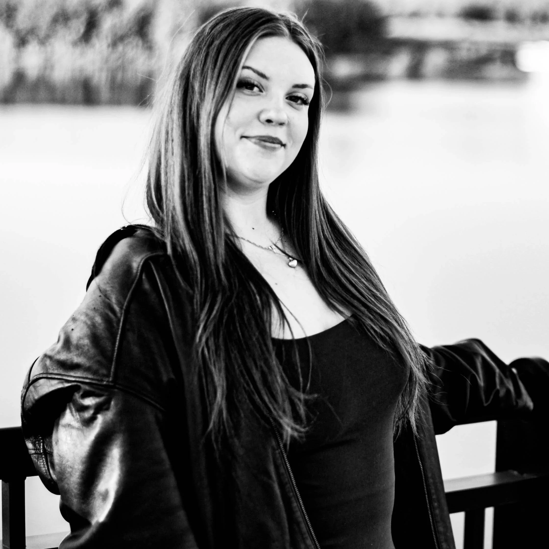 A woman with long hair wearing a leather jacket over a black top, sitting against a wooden railing outdoors, smiling slightly in a black-and-white photo.