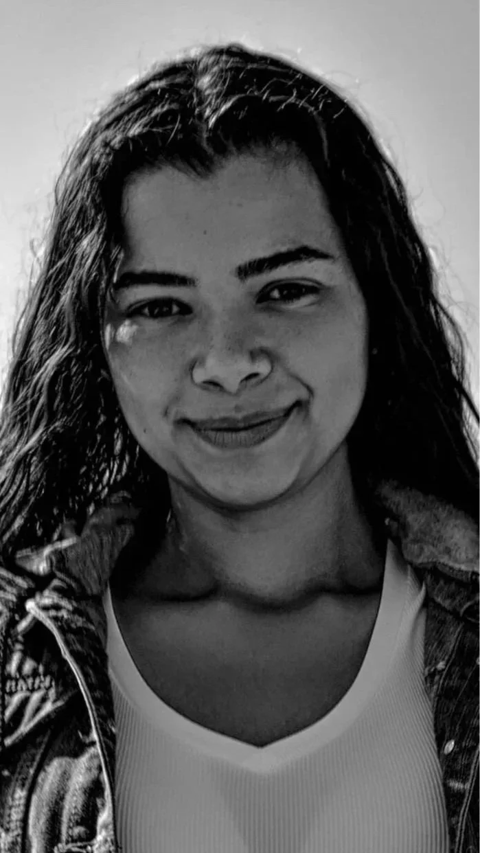 A black and white photo of a woman with long dark hair smiling, wearing a white tank top and a textured jacket.