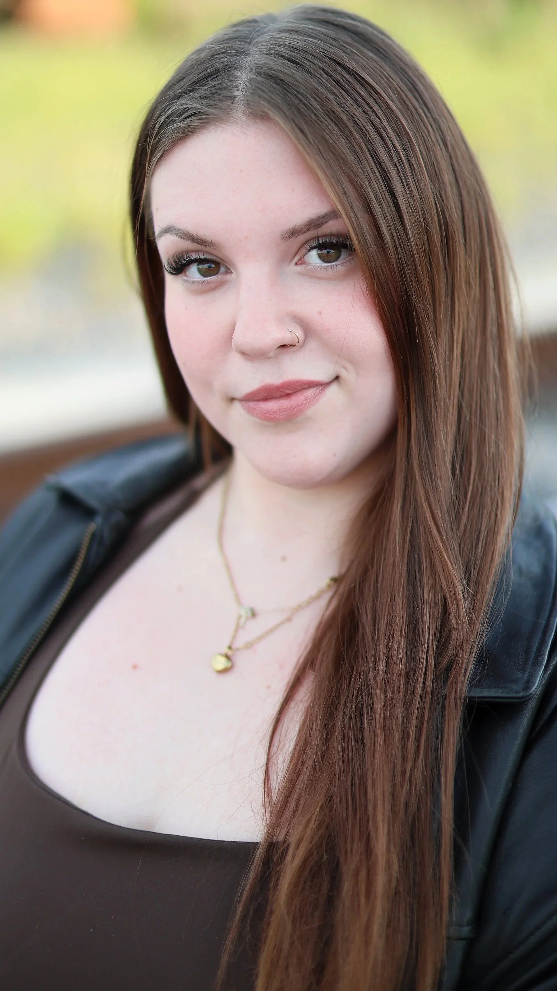 Close-up portrait of a young woman with long brown hair, wearing a black top, a leather jacket, a gold necklace, and small earrings, outdoors with a blurred background of greenery and a body of water.