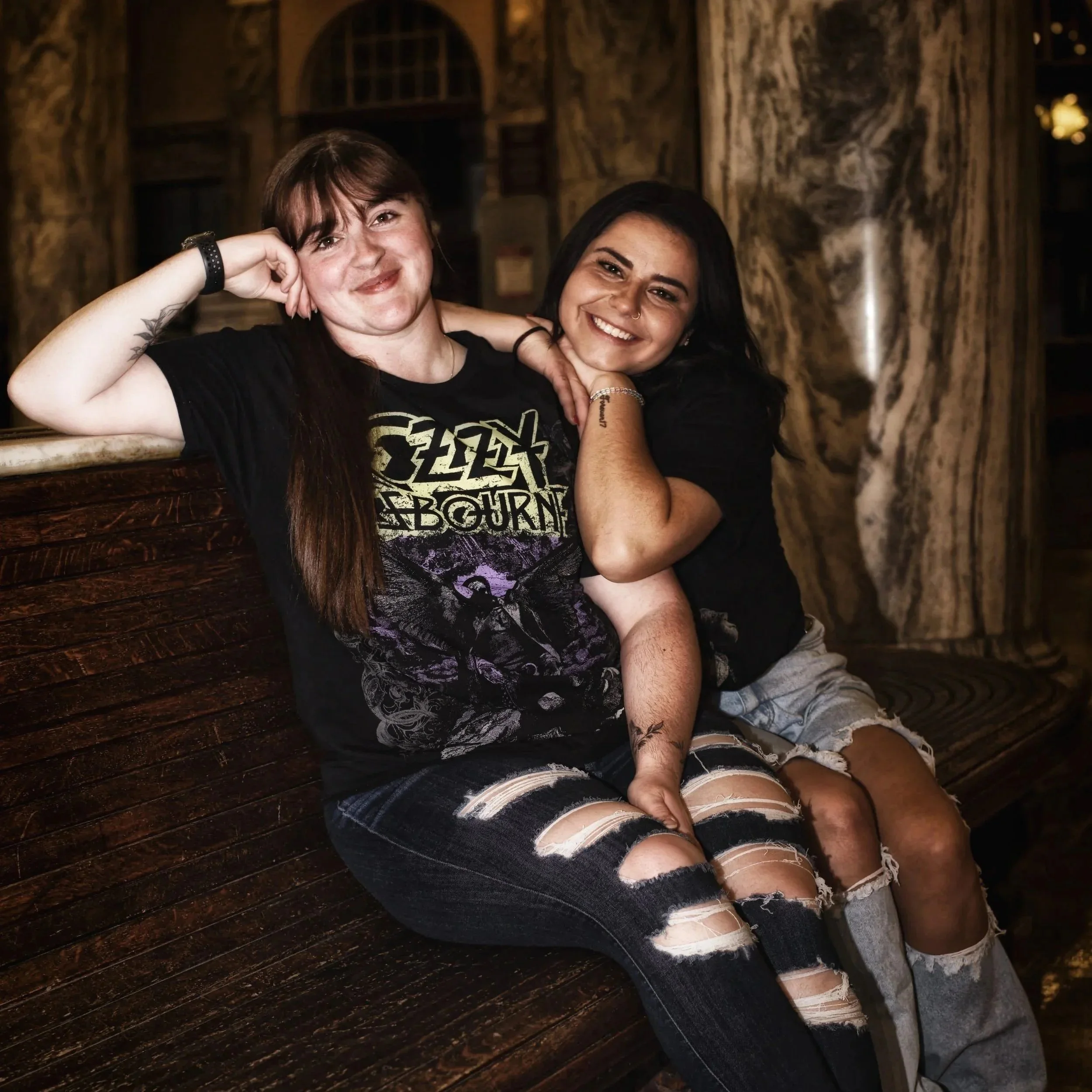 Two young women sitting close together on a wooden bench, smiling at the camera inside a warmly lit room with wooden walls and a marble column in the background.