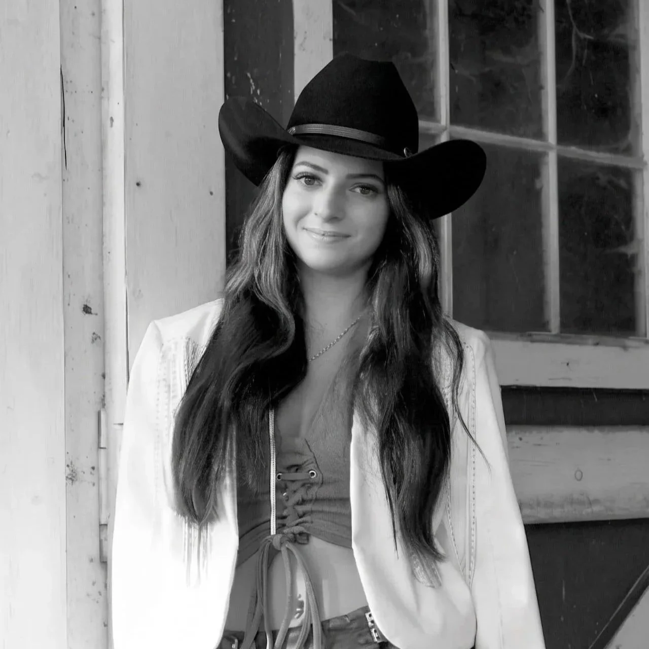 A woman with long dark hair wearing a black cowboy hat, a light jacket, a lace-up top, a short skirt, and cowboy boots, standing outdoors near a rustic wooden wall with a window.