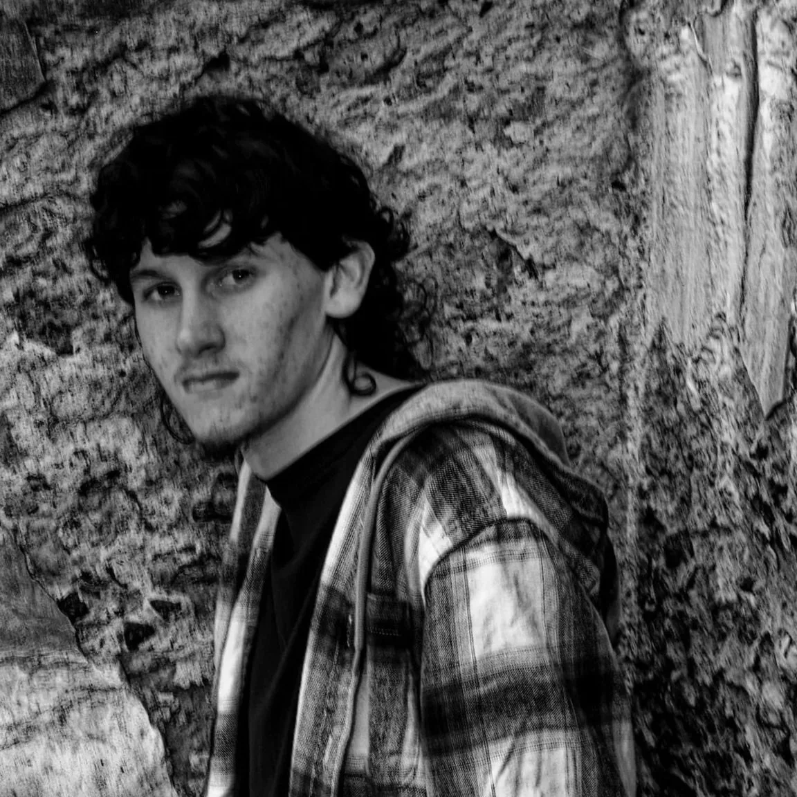 A young man with curly hair and a slight smile standing against a textured stone wall, wearing a plaid jacket over a dark shirt.