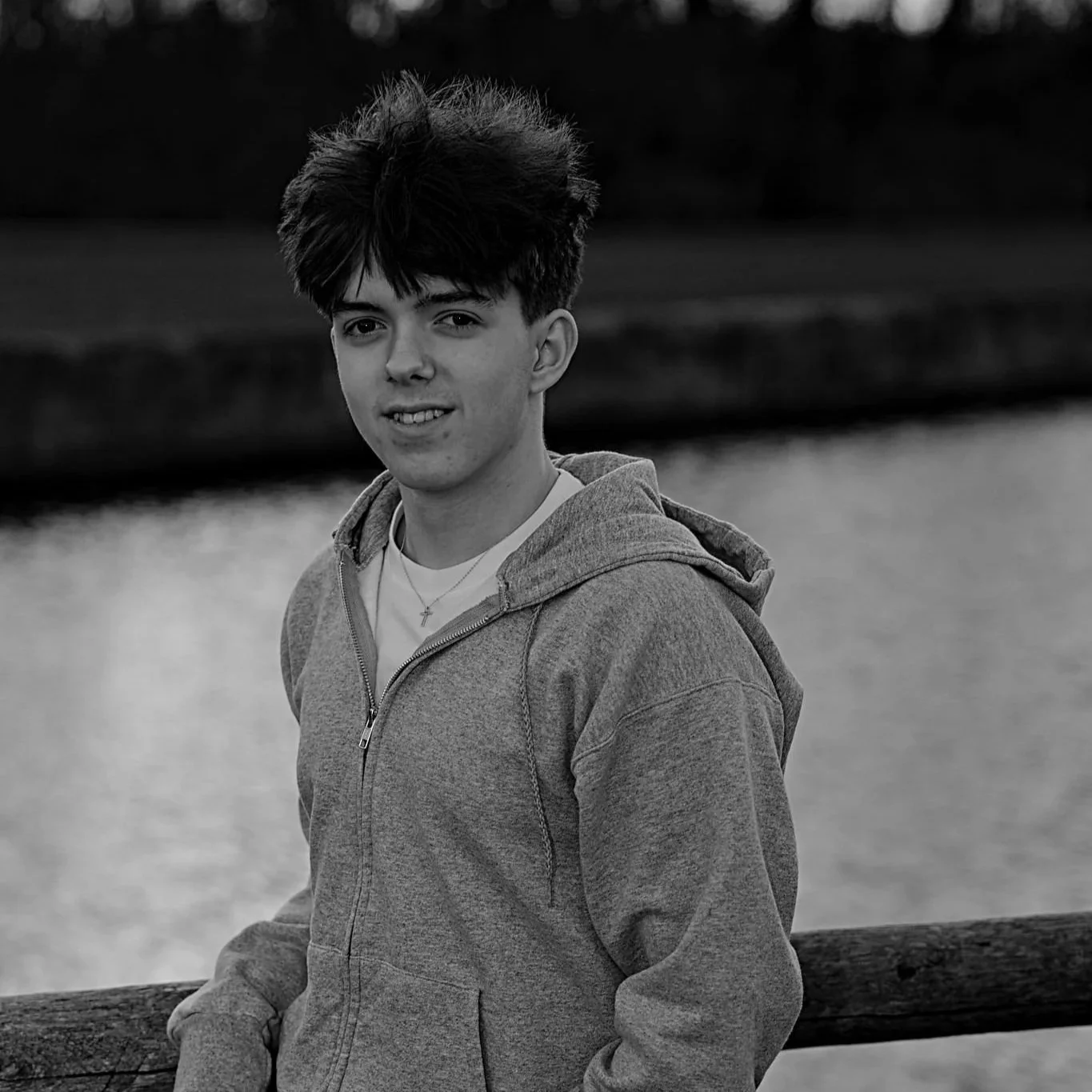 A young man with dark hair, wearing a hoodie and a necklace, standing outside near a body of water with trees in the background in a grayscale photo.