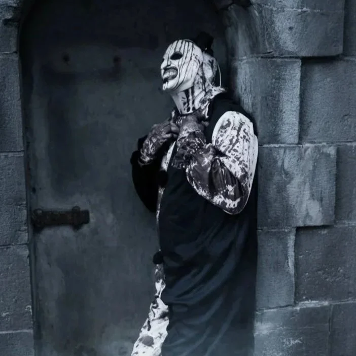 Person wearing a clown mask with black and white makeup, a black top hat, and gothic clothing, leaning against a stone wall near a metal door, surrounded by mist.