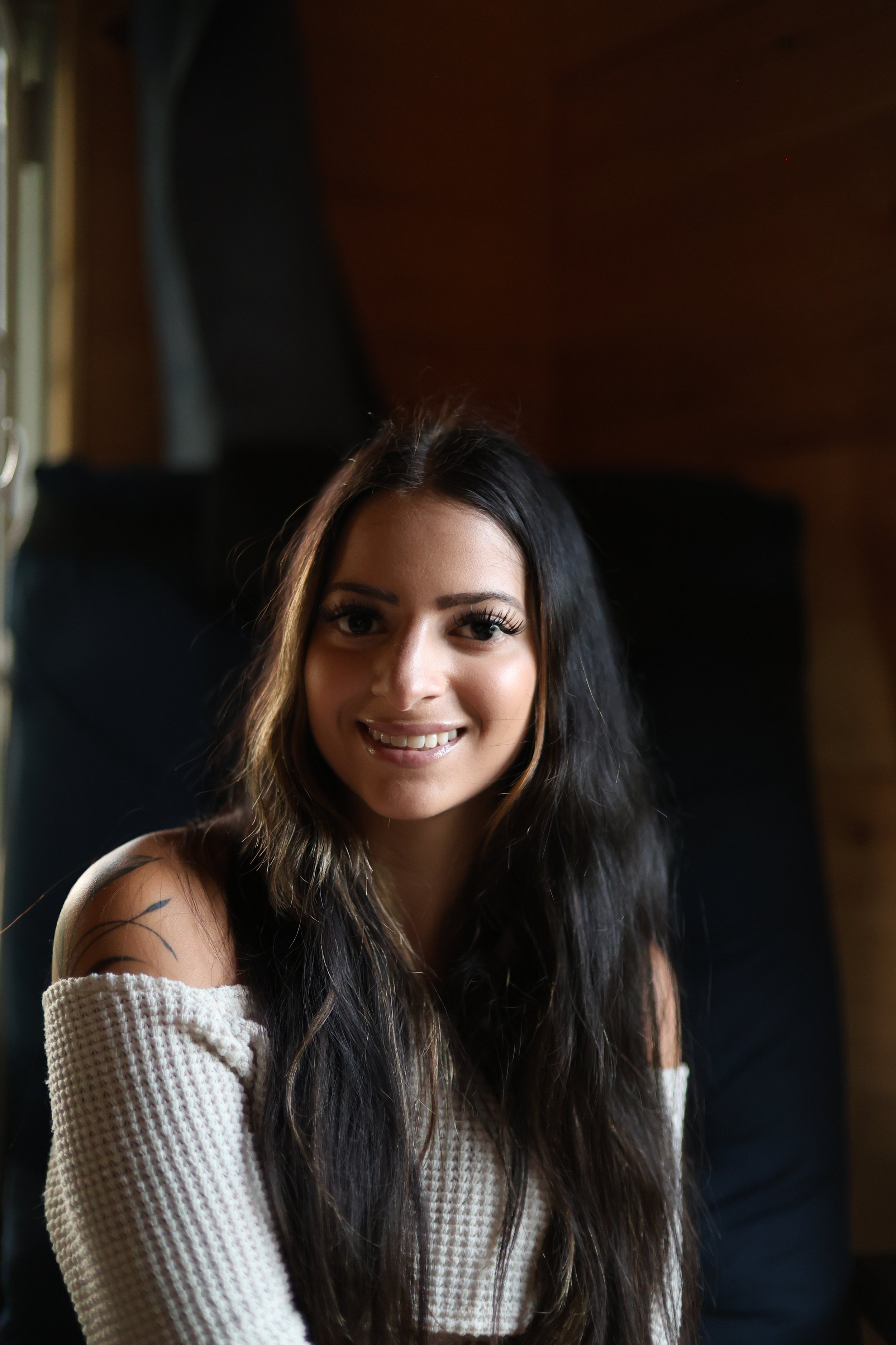 Close-up of a young woman with long dark hair, smiling, wearing a white off-shoulder sweater, with an arm tattoo visible, indoor setting with dark background.